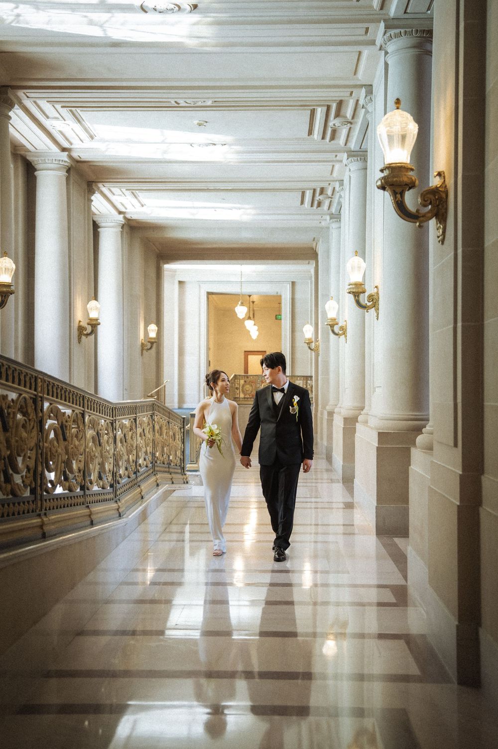 A couple walks down an elegant marble hallway with ornate columns and wall sconces in a grand historic building.