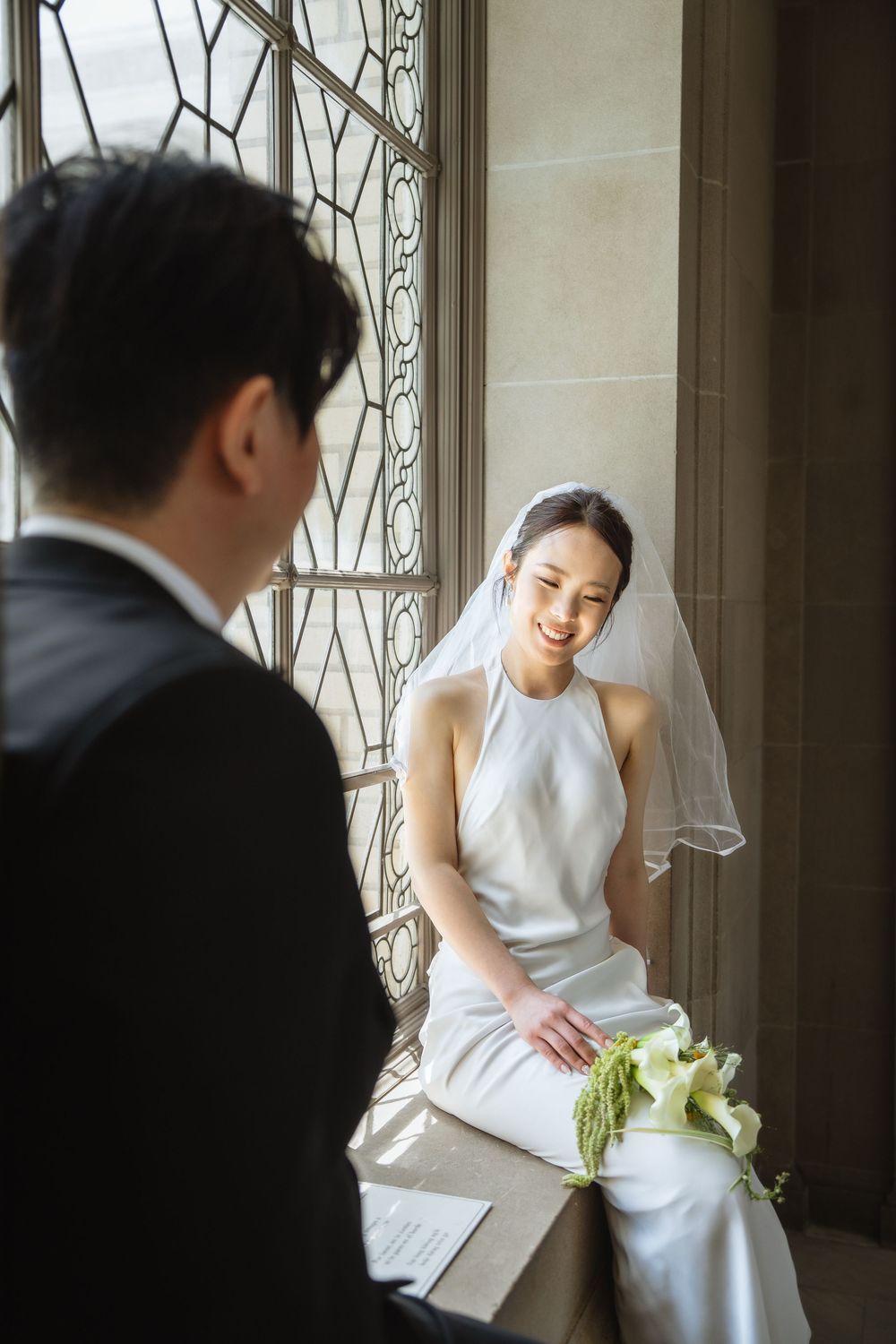 Bride in elegant white halter dress sits by ornate window holding calla lily bouquet while sharing a moment with groom.