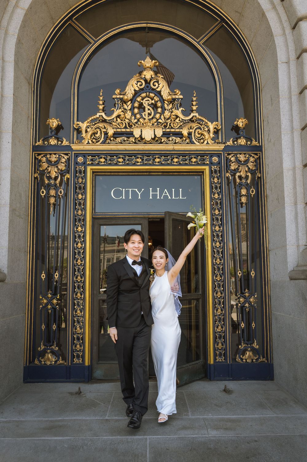 A couple celebrates their civil wedding ceremony in front of an ornate gold-trimmed entrance at City Hall.