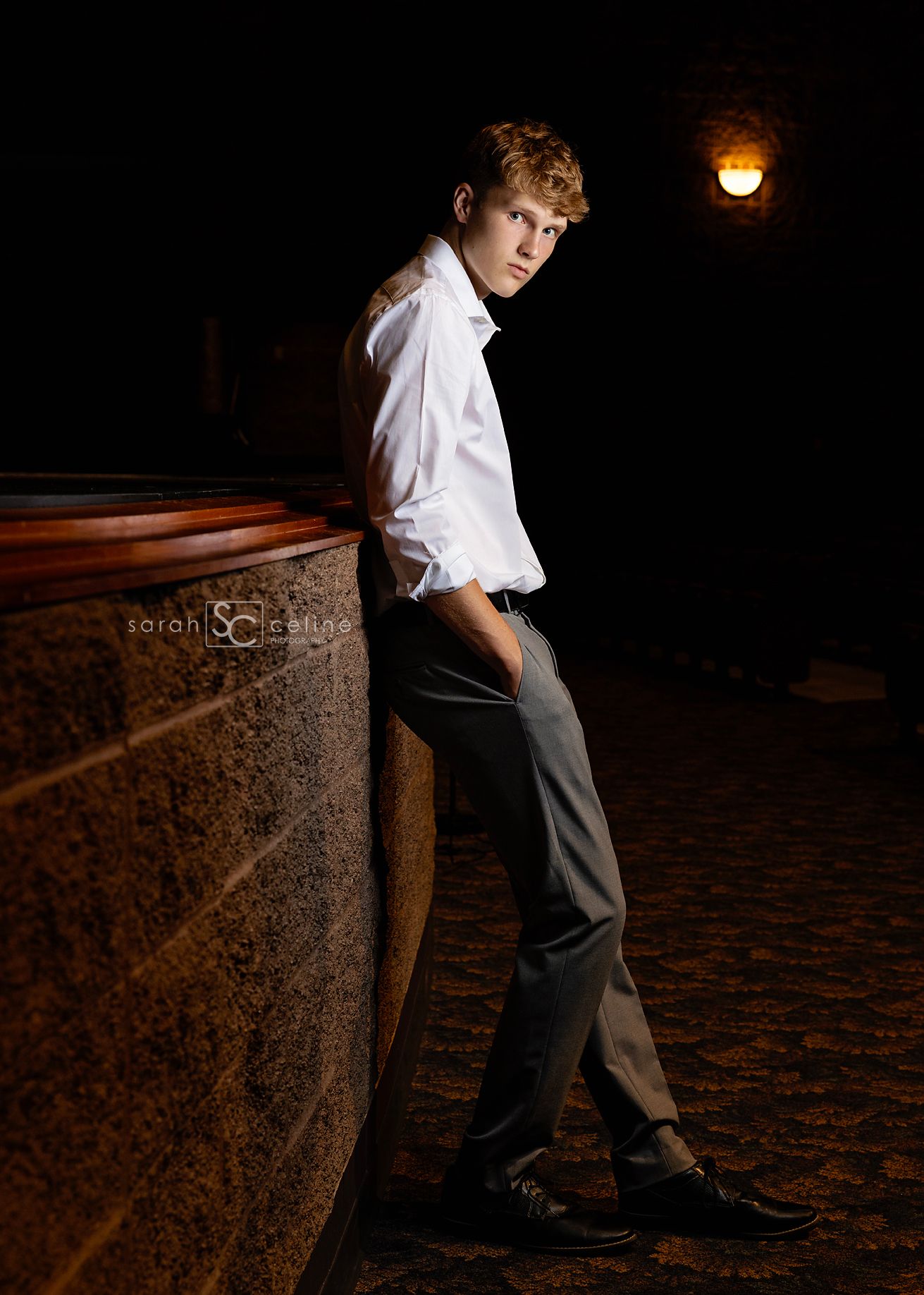 Young man in white shirt and dark pants poses against stone wall at night with vintage lamp glowing in background.