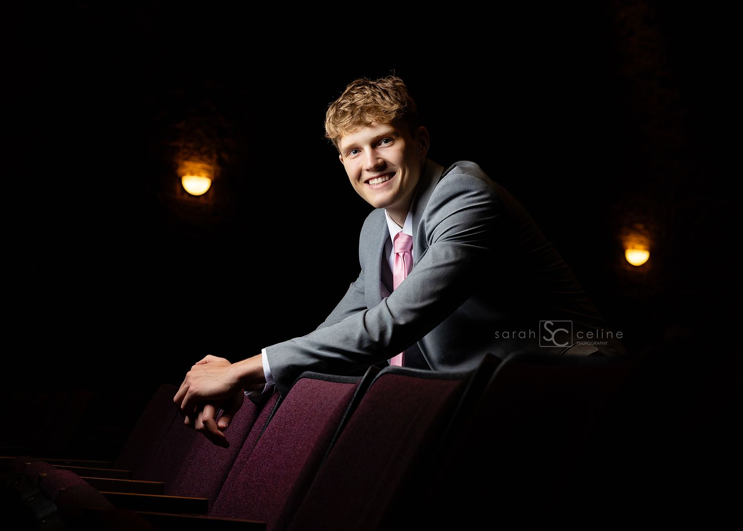 A person in a gray suit poses on dark red theatre seating with warm lighting in the background.