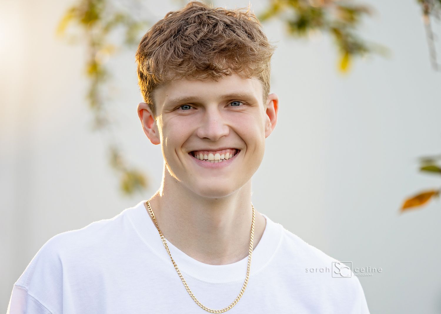 Happy portrait of someone wearing a white t-shirt and necklace against a light background with blurred foliage.