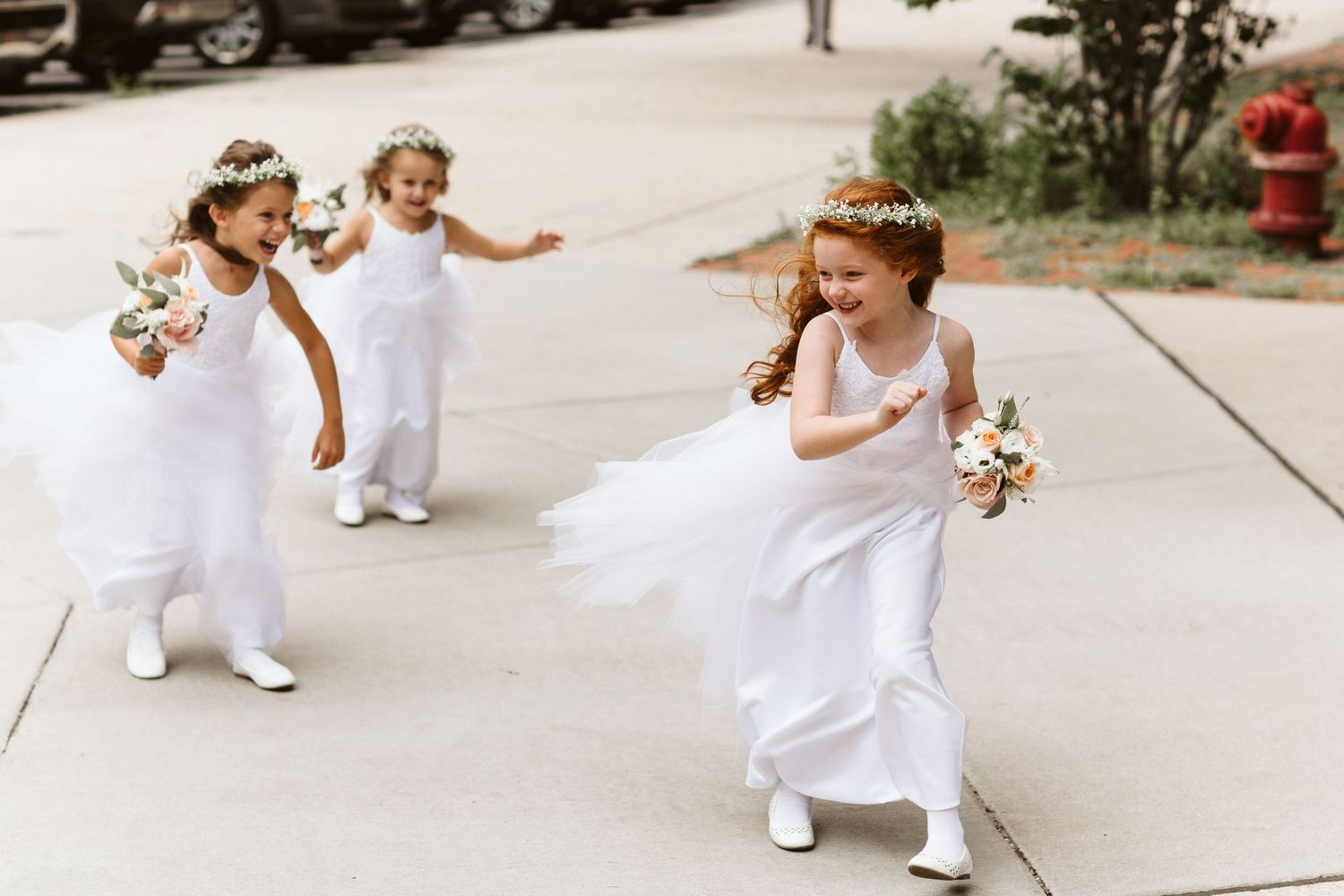 Young flower girls in white dresses walk down a sunlit path during a wedding ceremony.