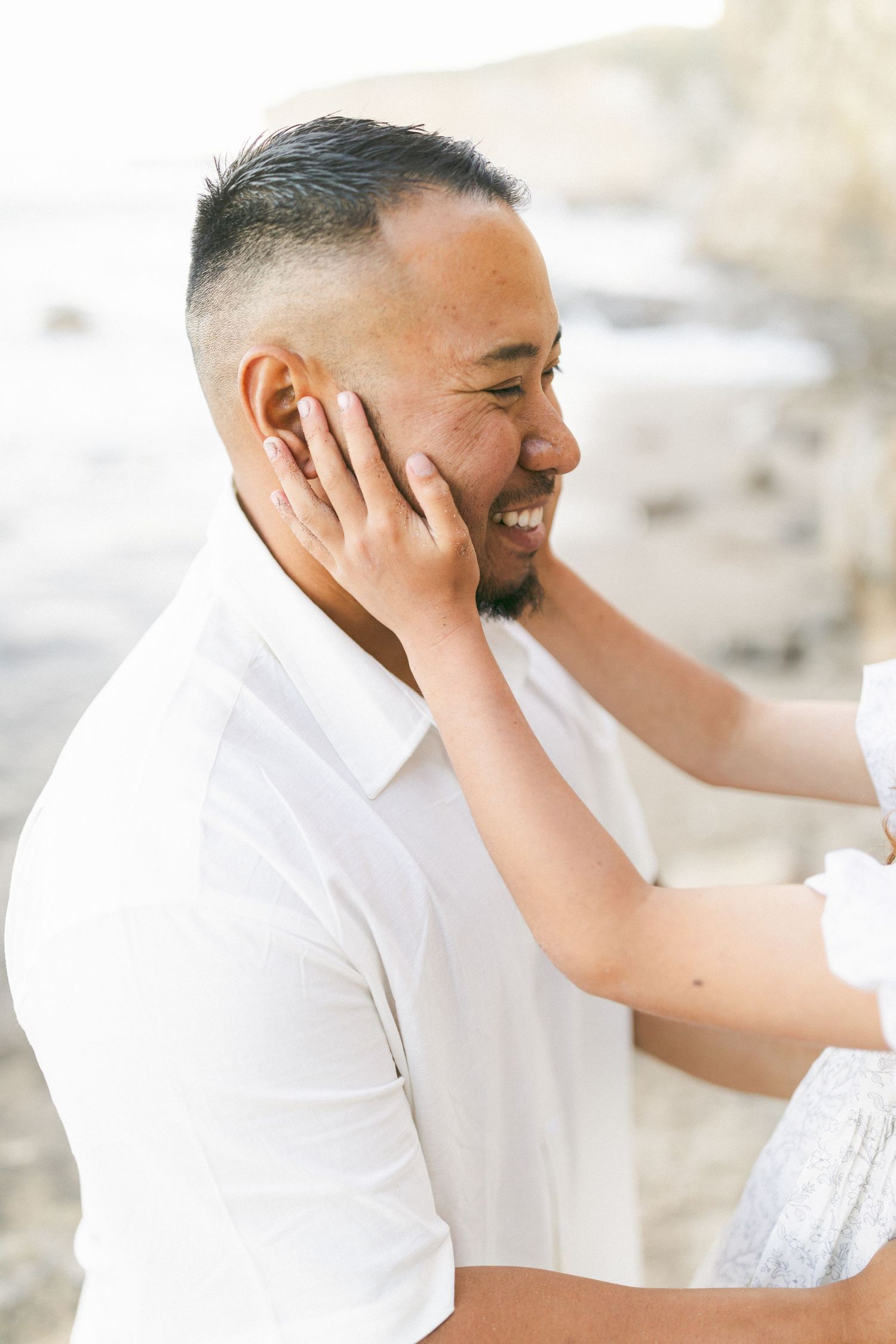 Someone wearing a white shirt smiles warmly while another person's hands gently touch their face in a tender moment captured by Chelsee Raw.