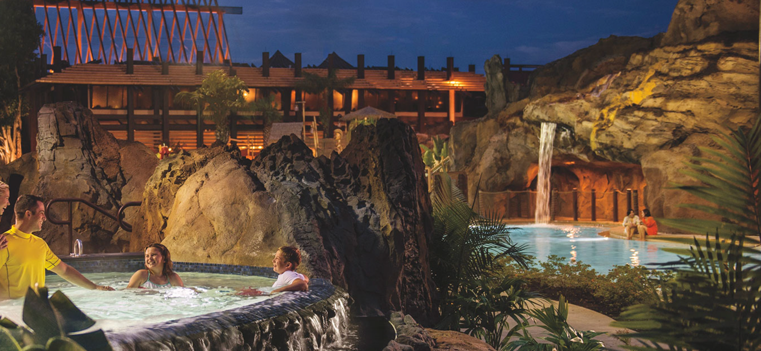 Evening view of resort exterior with illuminated pool area and tropical landscaping.