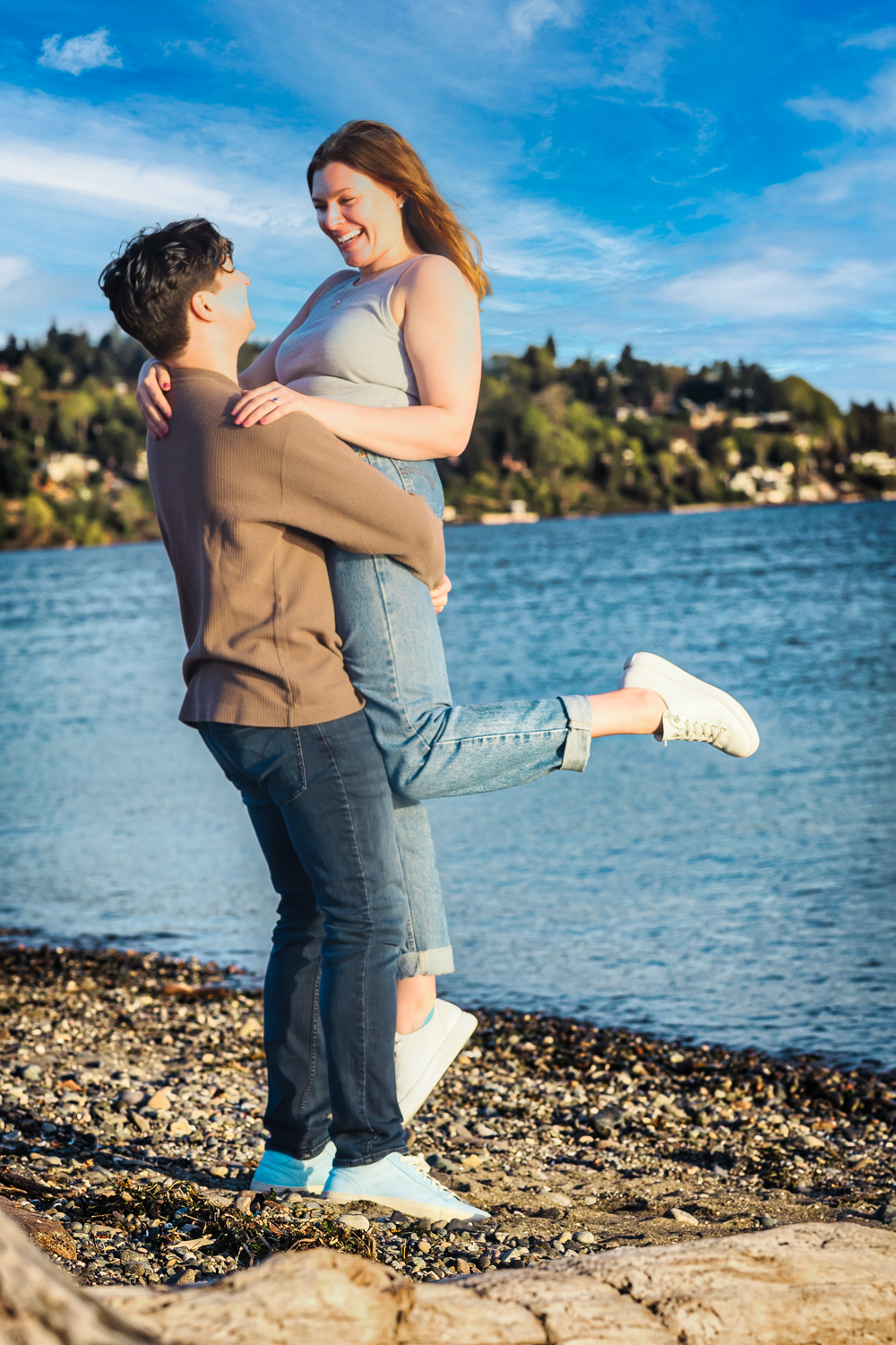 A couple embraces on a rocky beach at sunset with a lighthouse in the background.