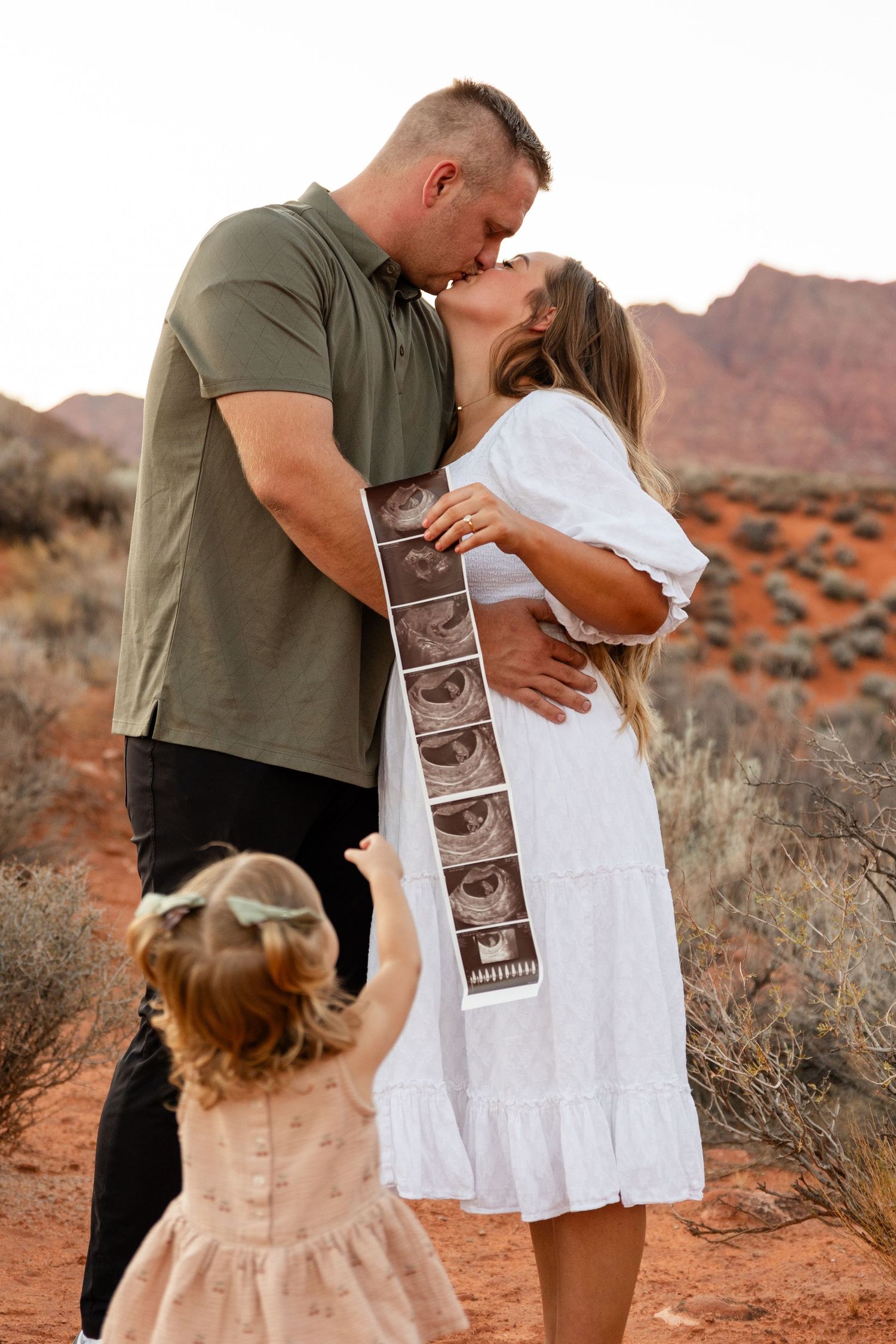 Southern Utah family photographer capturing mom and dad kissing while holding a sonogram as their 2-year-old reaches up to them in red rock
