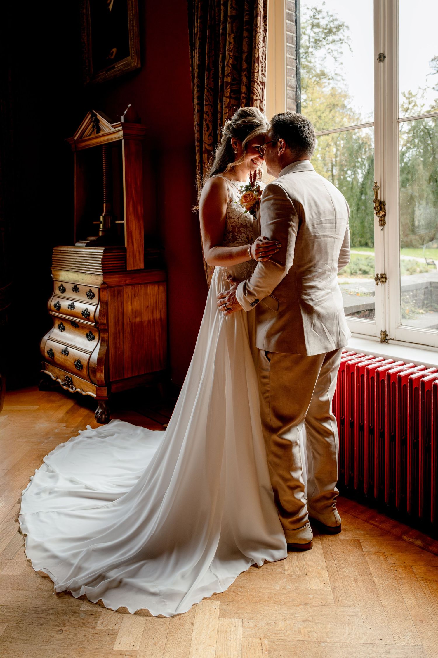 A romantic first look moment between newlyweds by a sunlit window in a rustic indoor venue with wooden floors in Geldrop.
