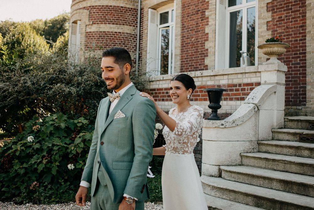 Bride in floral lace gown touches groom's shoulder in sage suit during first look at elegant brick estate.