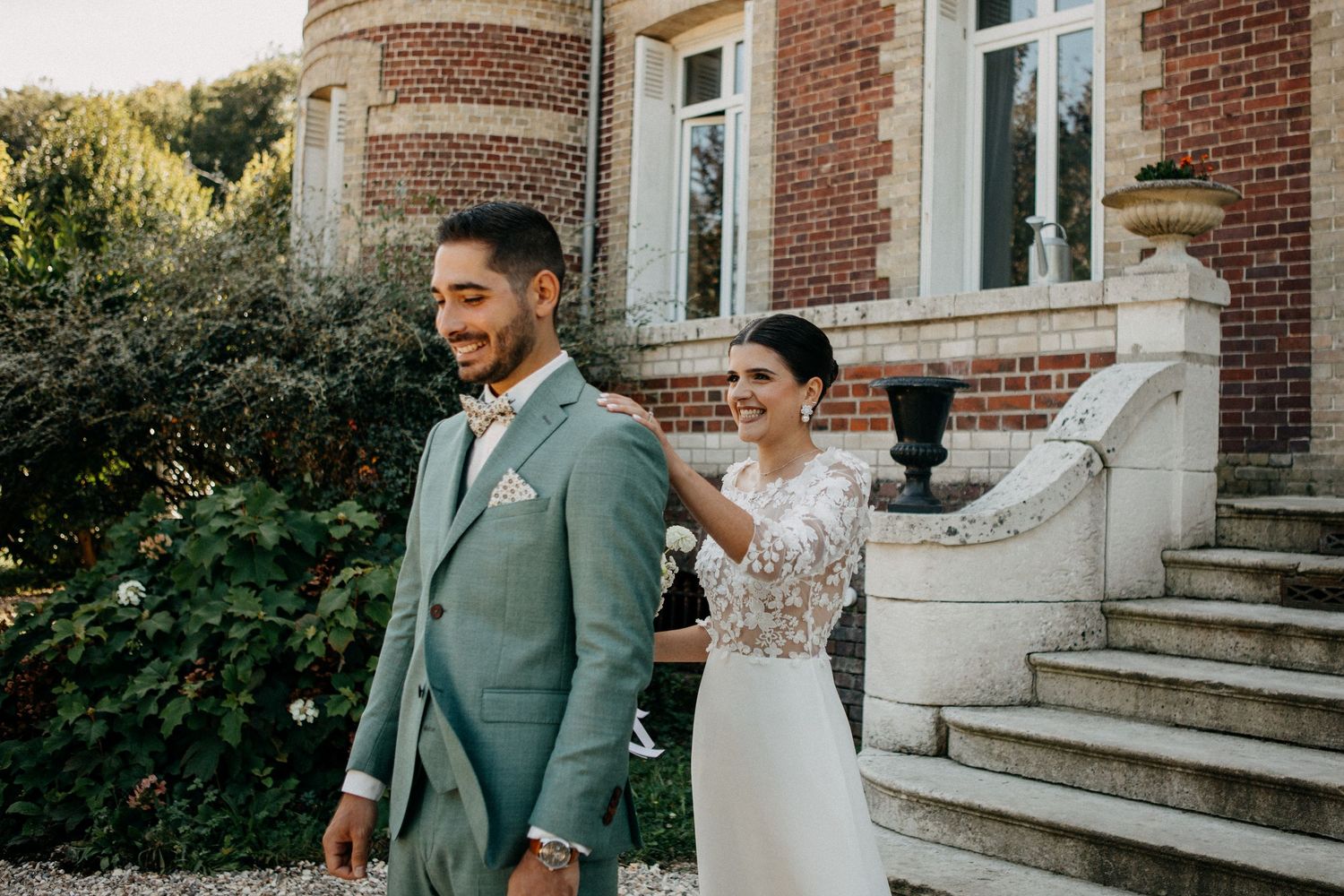 Bride in floral lace gown touches groom's shoulder in sage suit during first look at elegant brick estate.