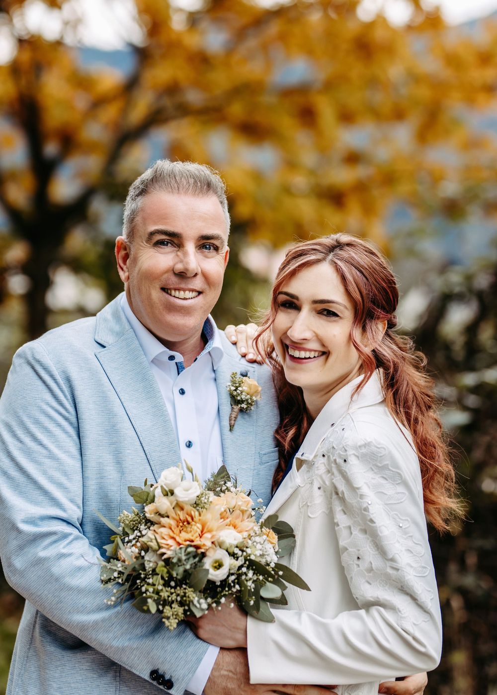 A happy couple poses outdoors on their wedding day with autumn foliage in the background and a peach-colored bouquet.