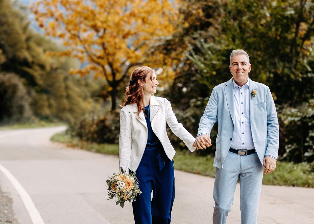 A couple walks together on a winding road surrounded by vibrant autumn foliage in golden hues.