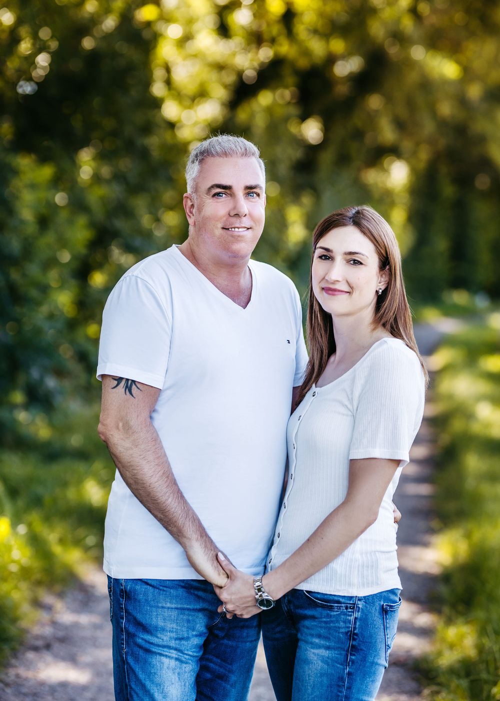 A couple in matching white t-shirts and jeans embracing outdoors with sunlit trees in the background.