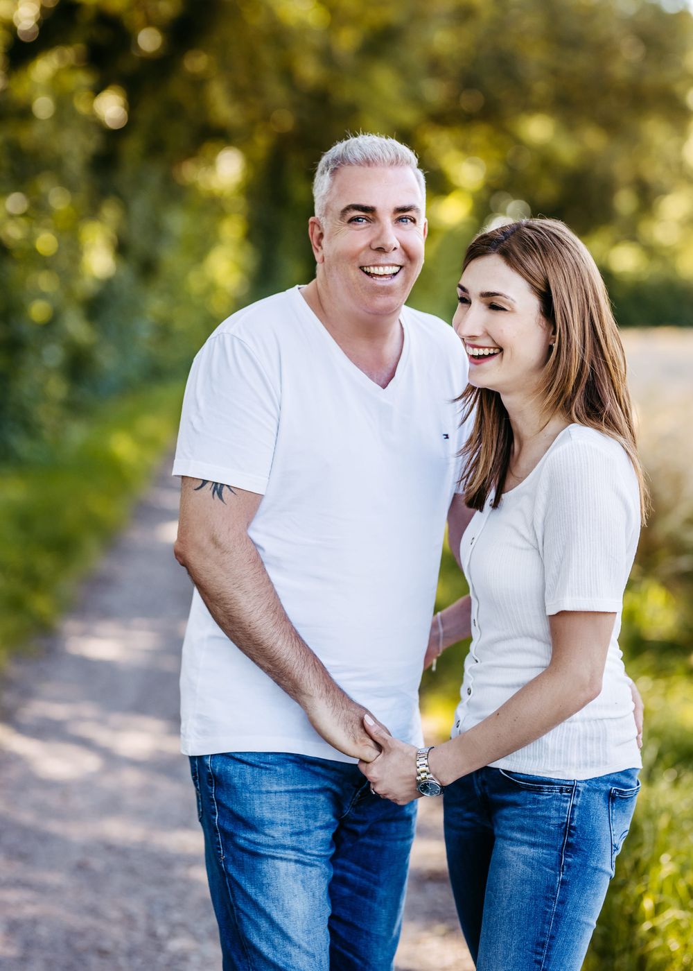 A couple wearing matching white t-shirts and jeans laugh together on a sunny outdoor path with bokeh lighting.