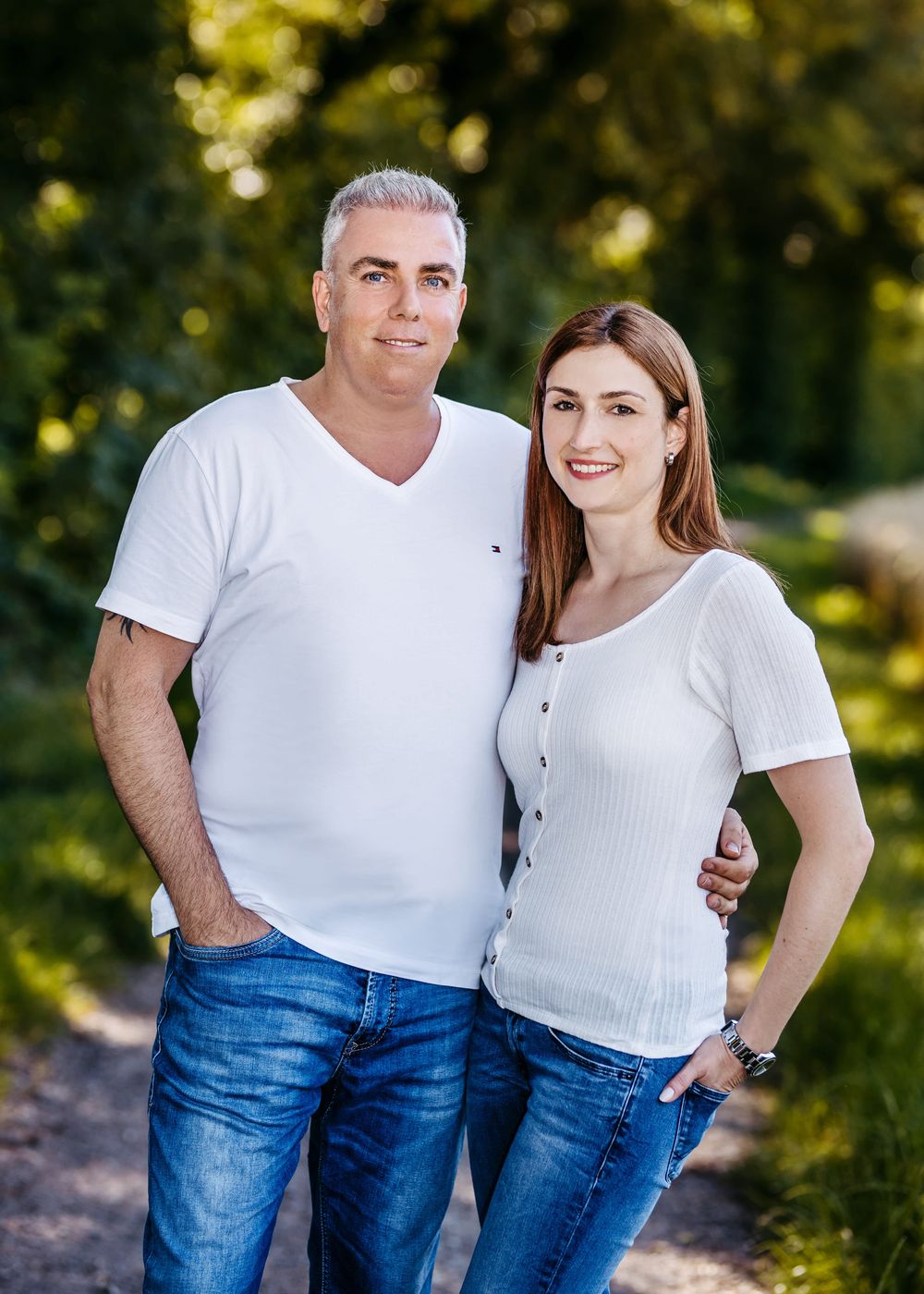 A couple wearing matching white t-shirts and blue jeans pose together outdoors in a natural setting with bokeh background.