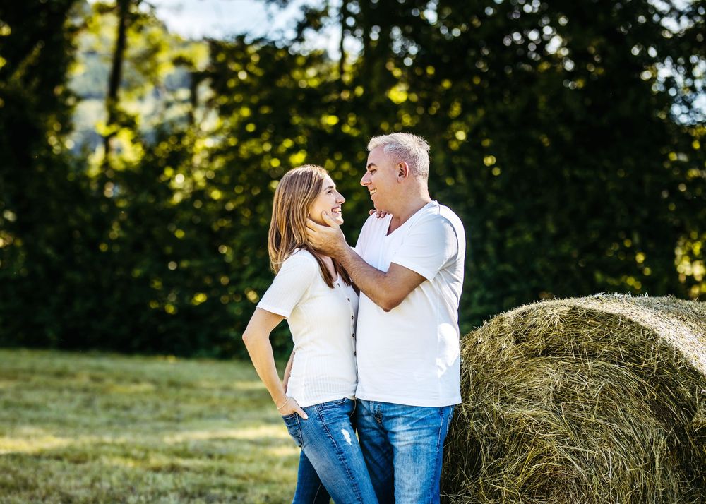 Romantic couple embracing in a summer field with hay bales and trees in the background.