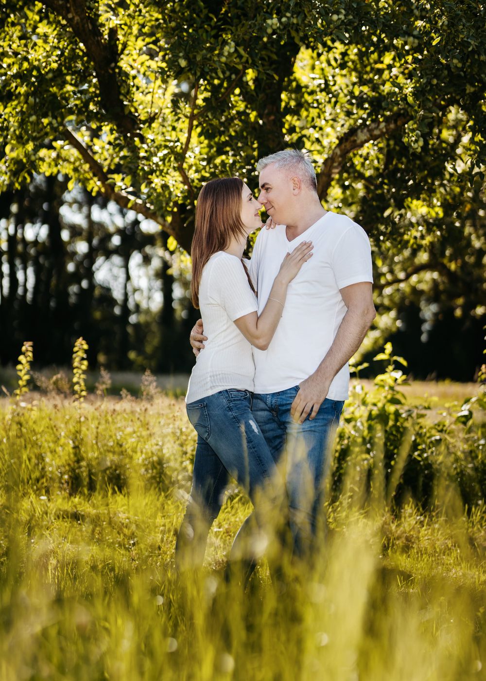 A couple embraces in a sunlit meadow with golden light filtering through trees in the background.