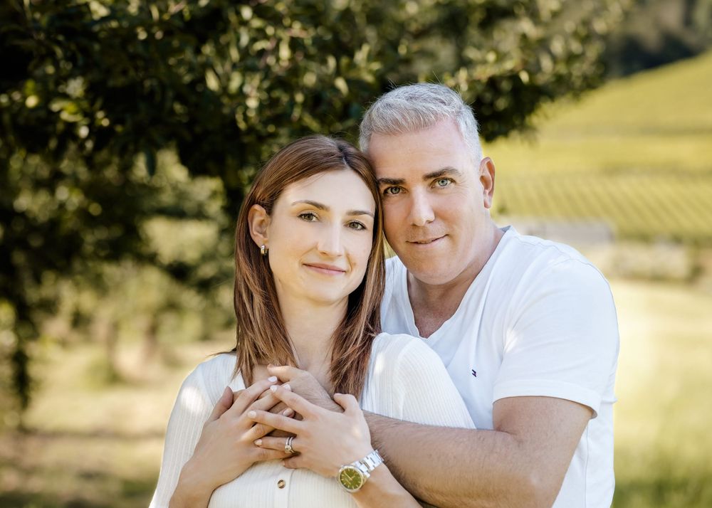 A couple in white outfits shares tender moments during an outdoor photoshoot in a sunny natural setting.