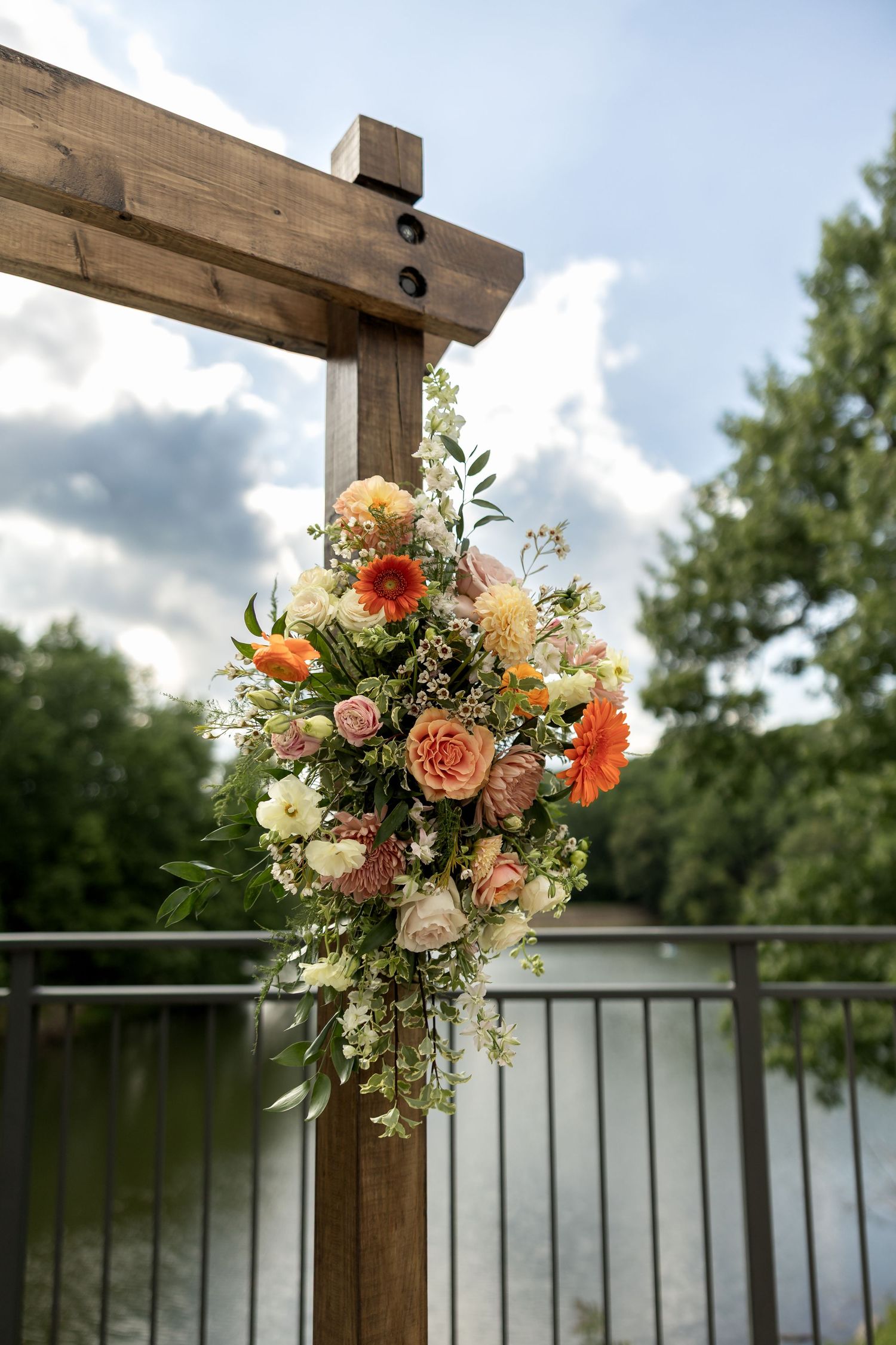 Orange and pink floral arrangement adorns a wooden arch post overlooking a serene lake.