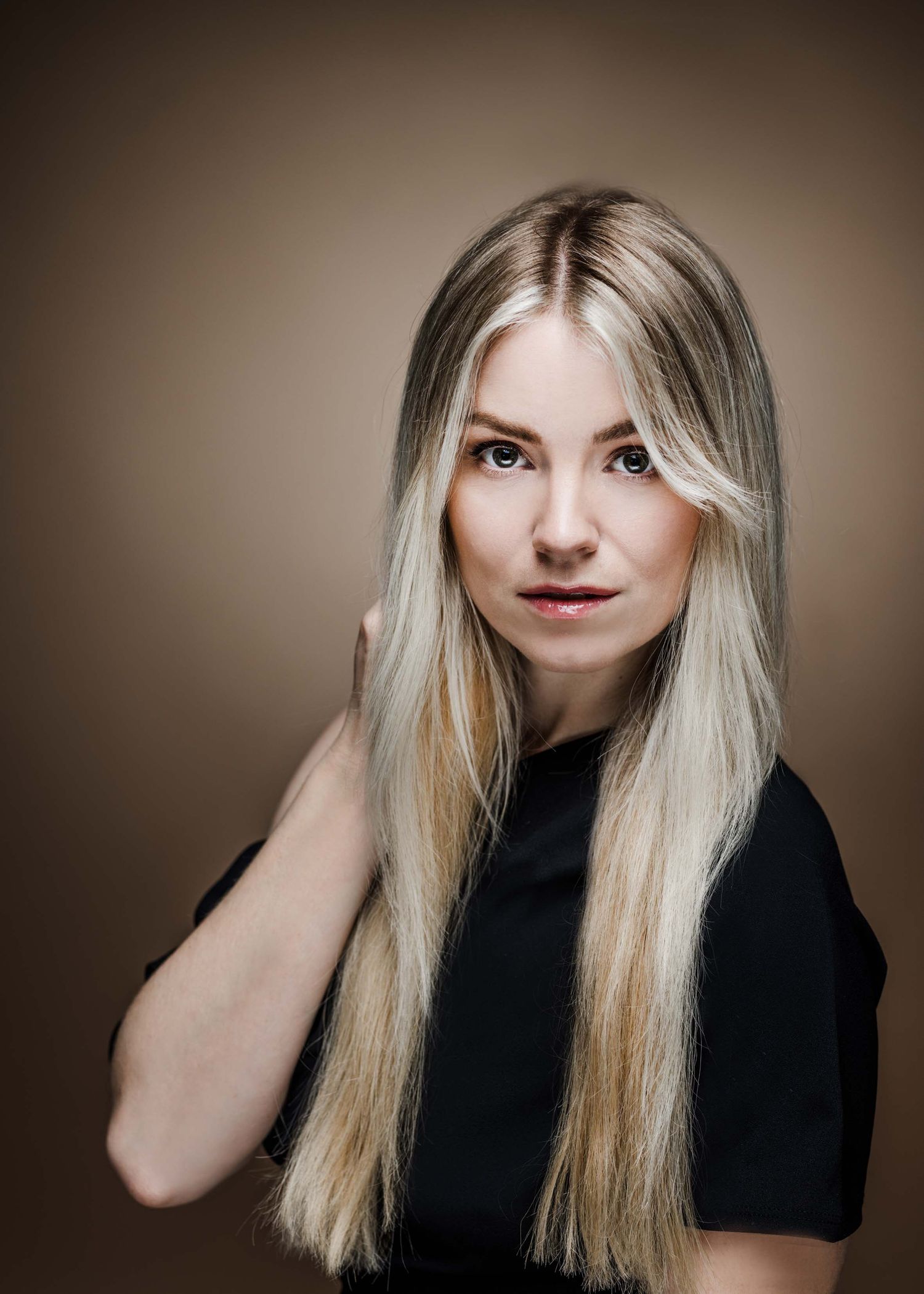 Studio portrait of a person with long blonde hair wearing a black top against a brown background.