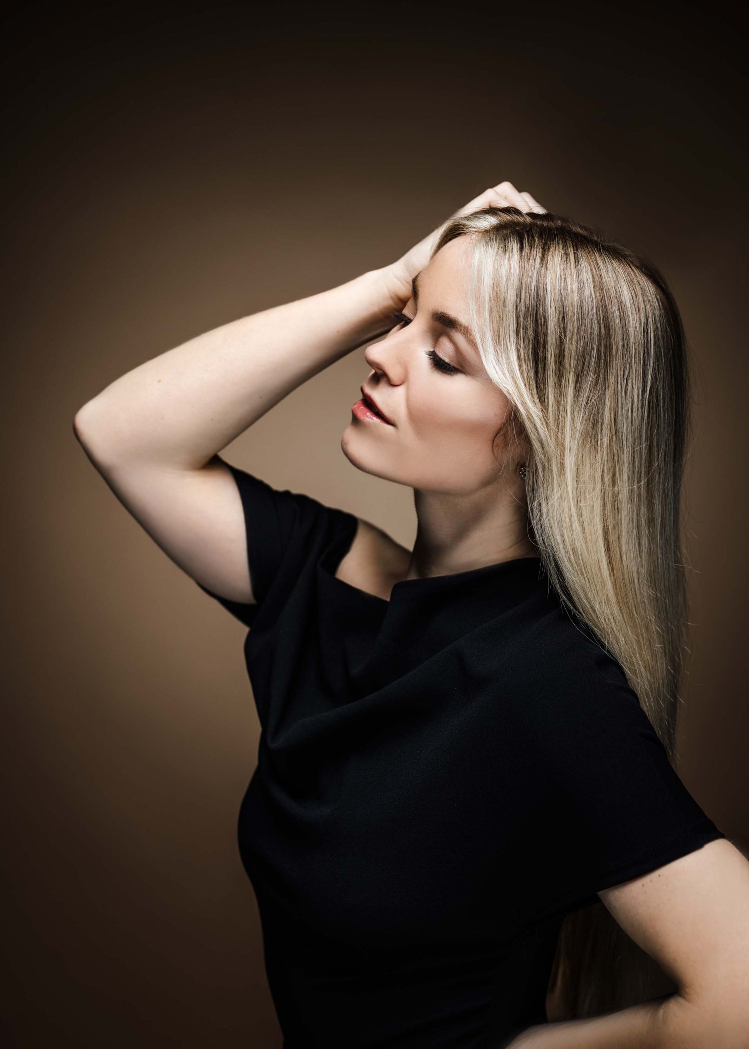 Elegant portrait of a person with long blonde hair wearing black against a dark brown backdrop.