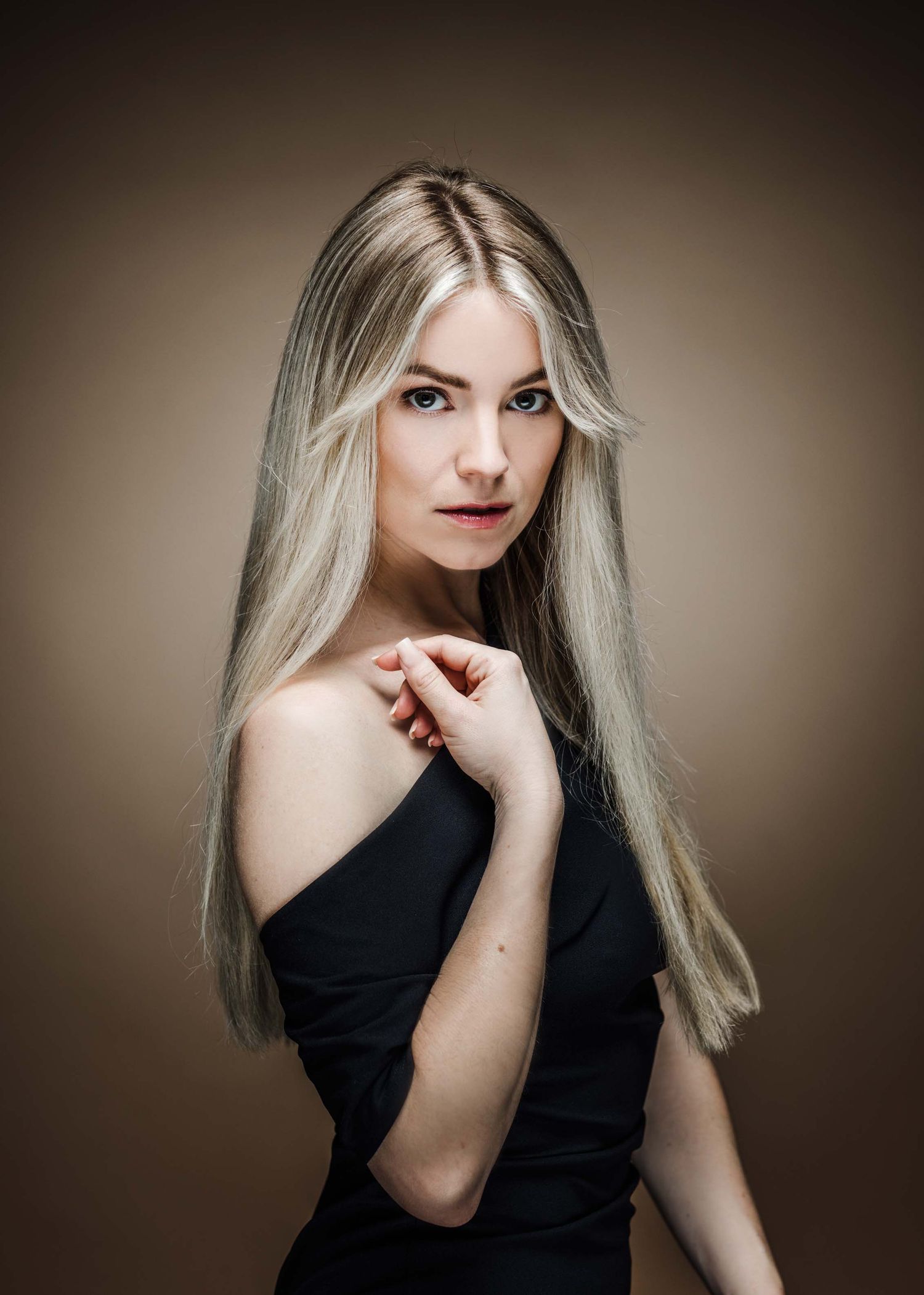 Studio portrait with long platinum blonde hair wearing a black off-shoulder top against brown backdrop.
