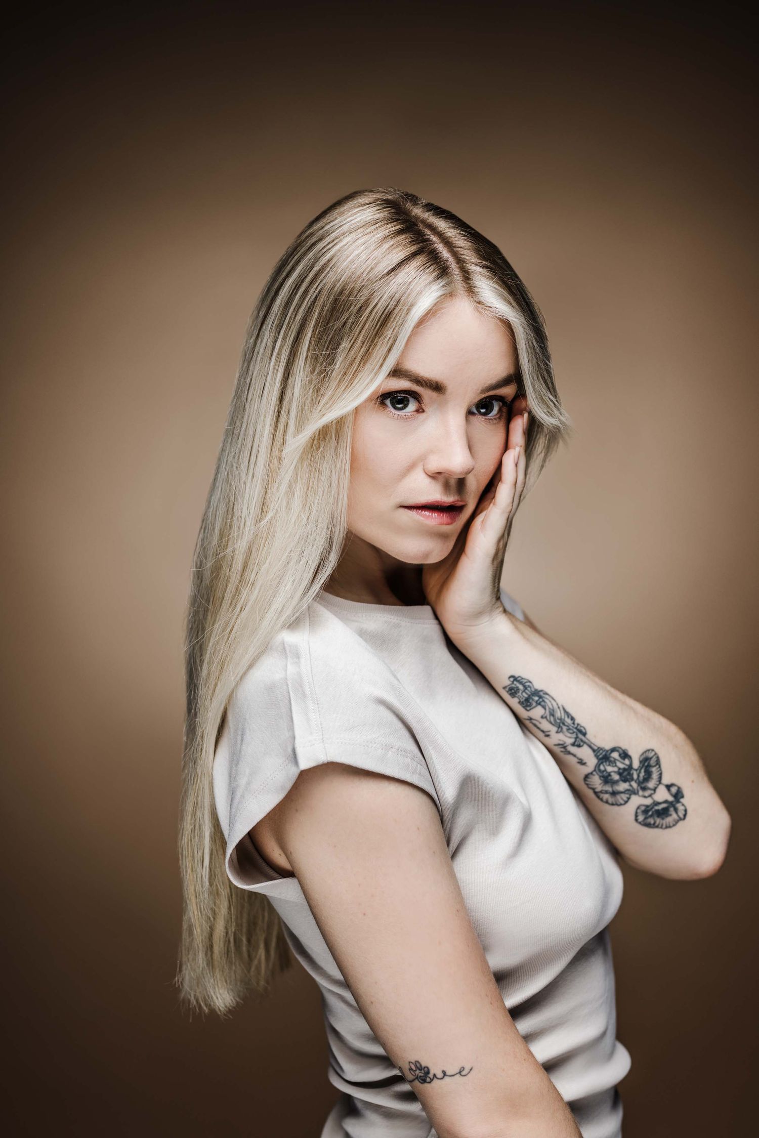 Studio portrait of a person with long platinum blonde hair wearing a white top and showing arm tattoos against brown backdrop.