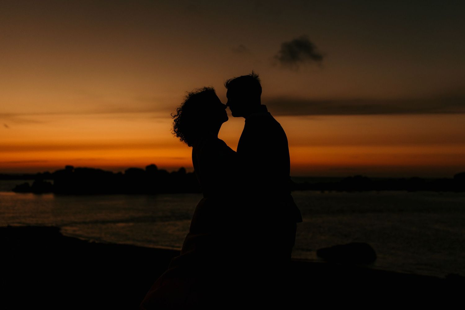 Silhouetted couple embracing against dramatic orange sunset over water.