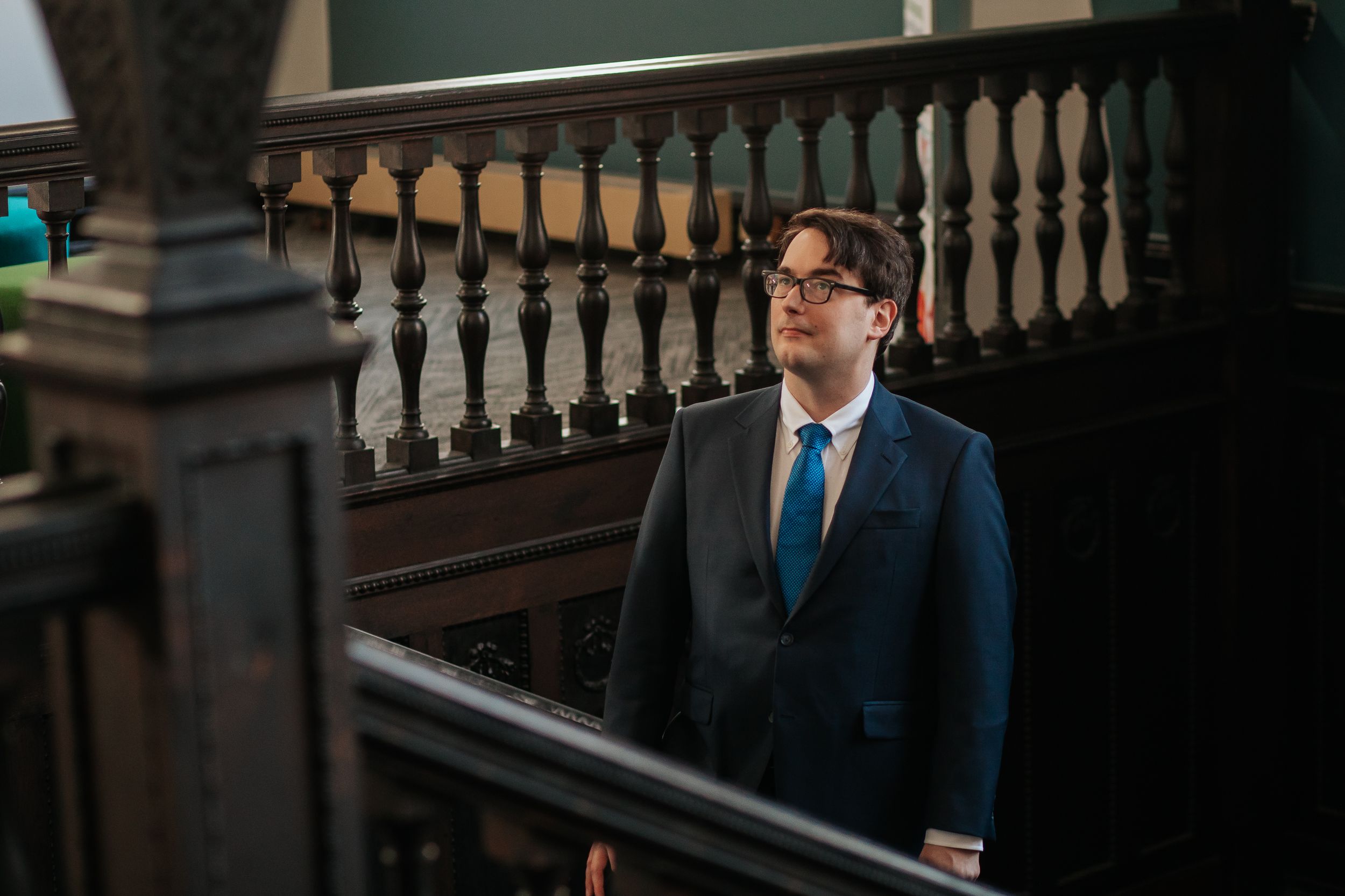 A person in a dark suit and tie stands thoughtfully against an ornate wooden railing in an elegant interior setting.