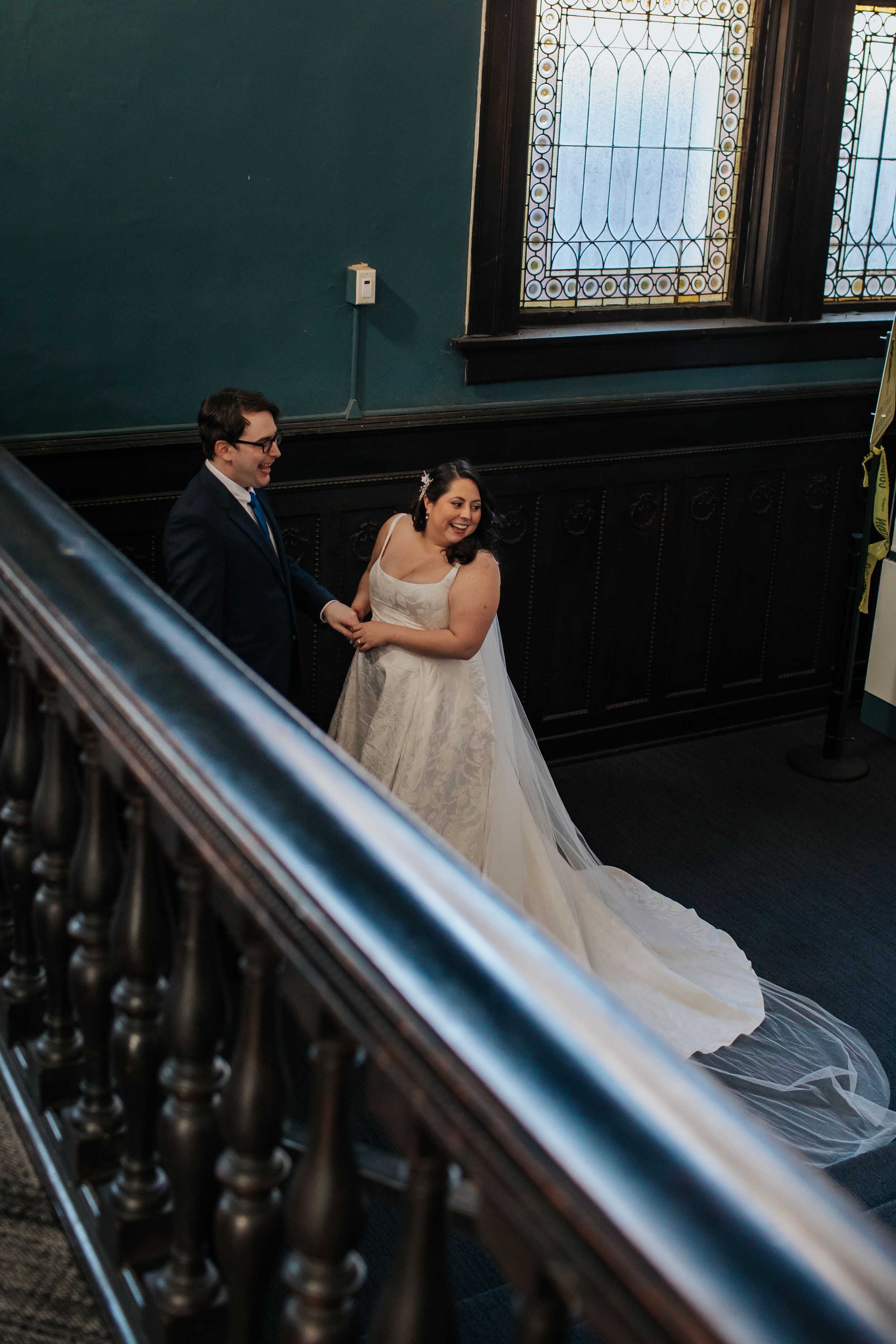 A couple stands together on an elegant staircase with ornate railings and stained glass windows in a historic building.