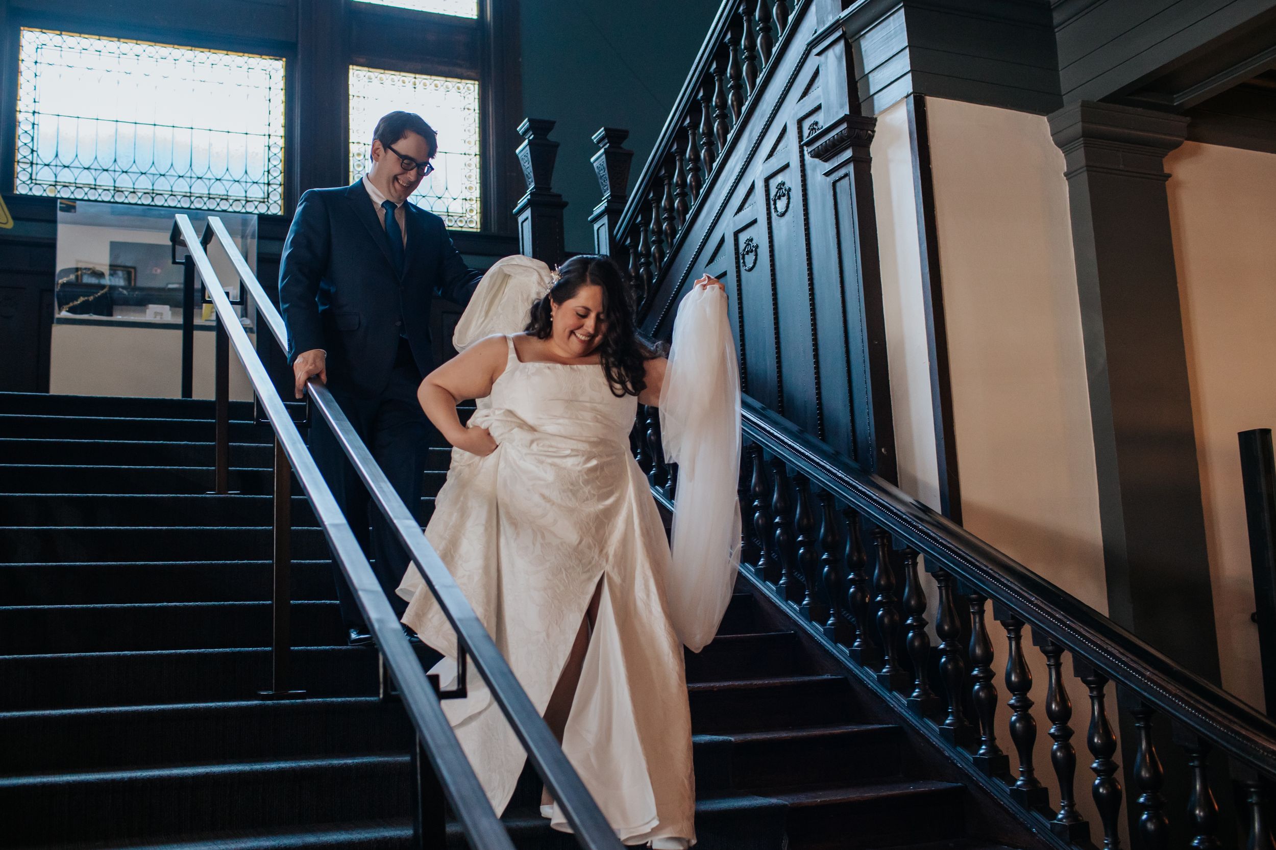 A couple in formal attire descends an elegant staircase with ornate black railings inside a historic building.
