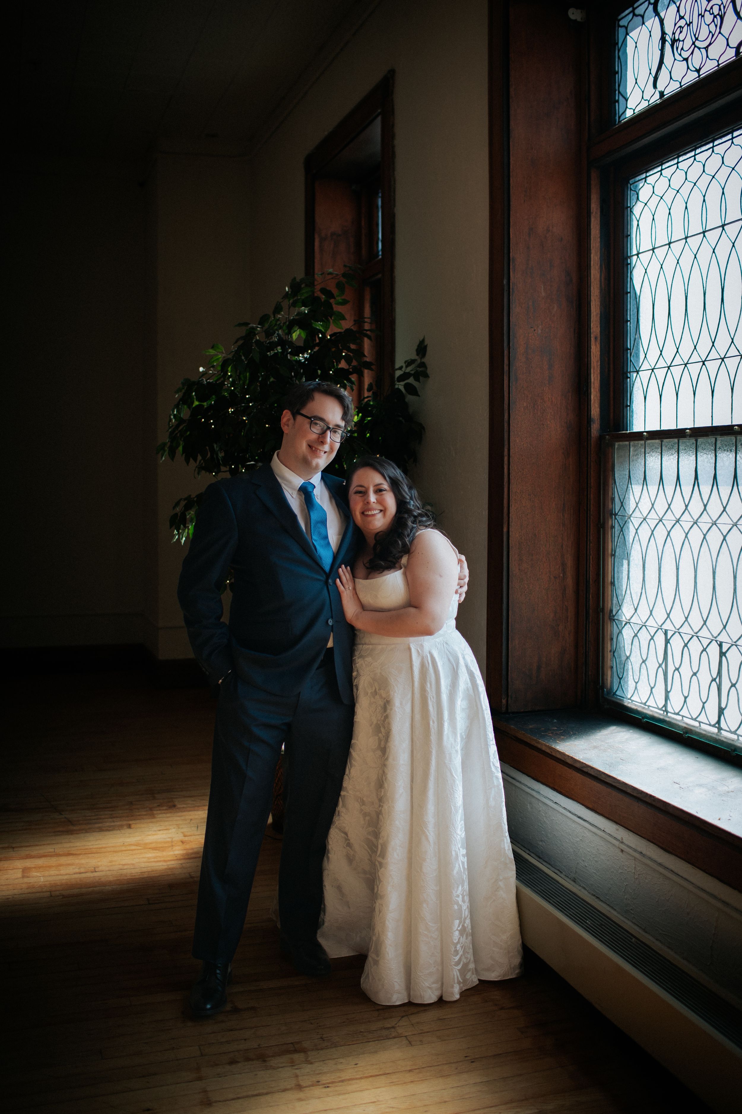 A couple in wedding attire stands together by a window in a dimly lit room with wooden floors.