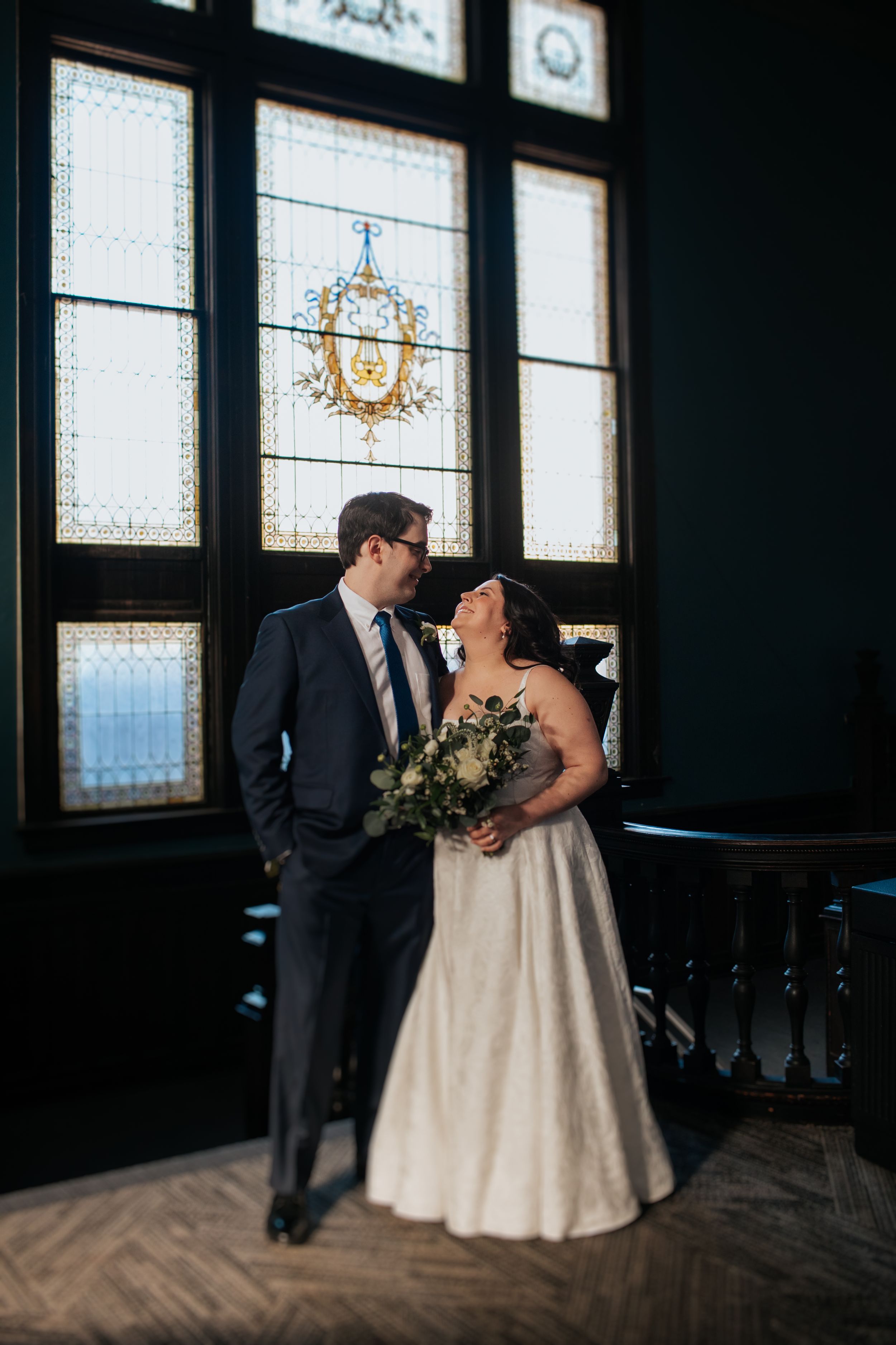 A couple in wedding attire share an intimate moment in front of decorative windows in an elegant dark room.