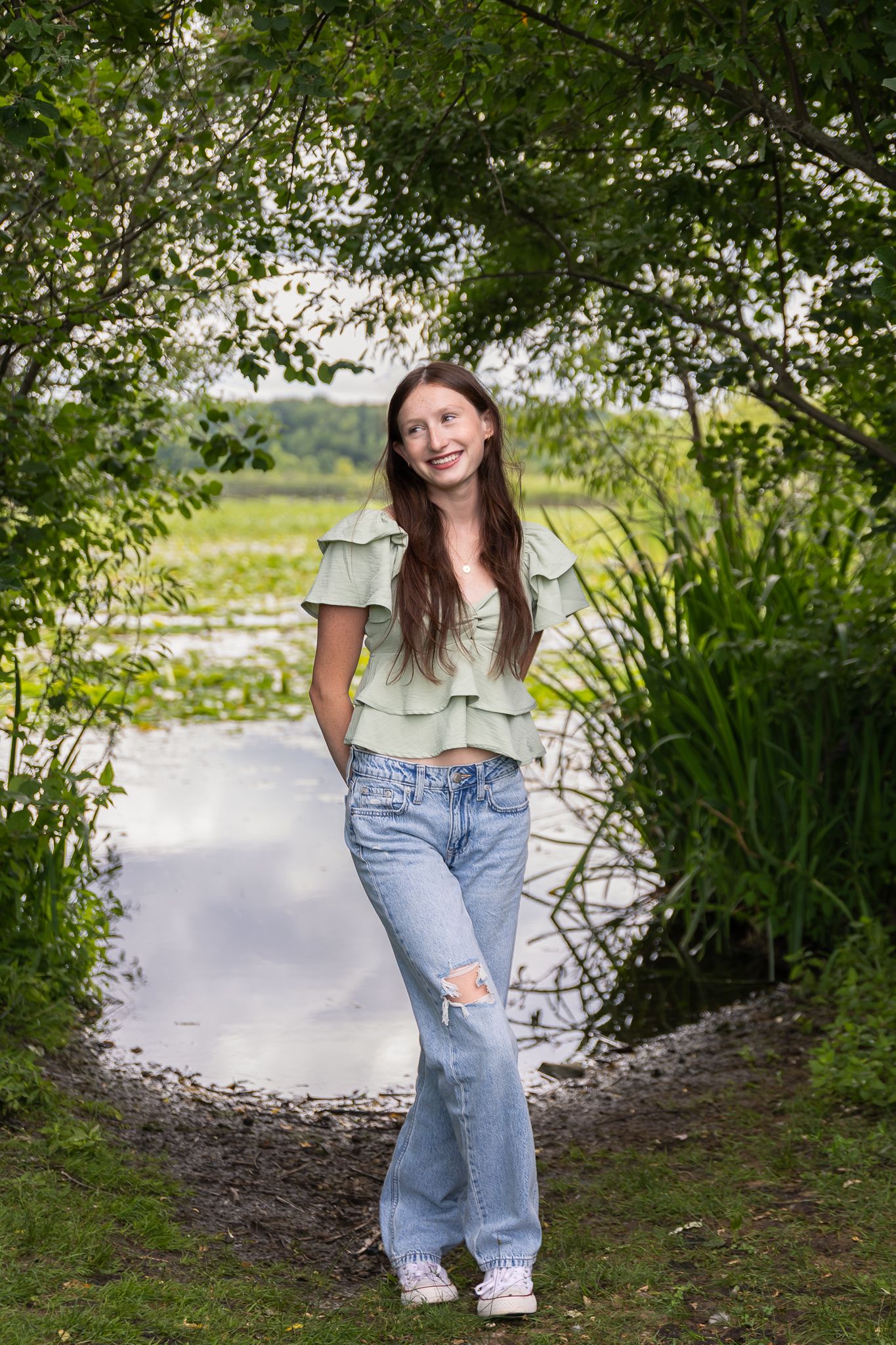 High school senior girl poses in front of trees and a wetland at Crosswinds Marsh in Wayne County, MI. She is smiling and dressed casual.