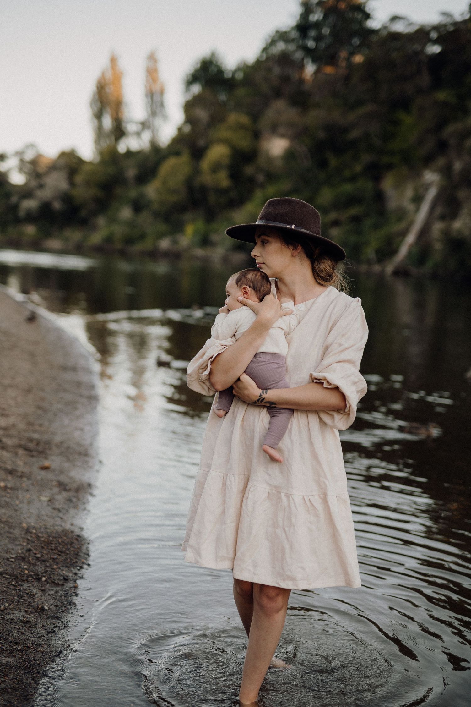 A woman in a white dress and wide-brim hat wades in a river while cradling a newborn baby.