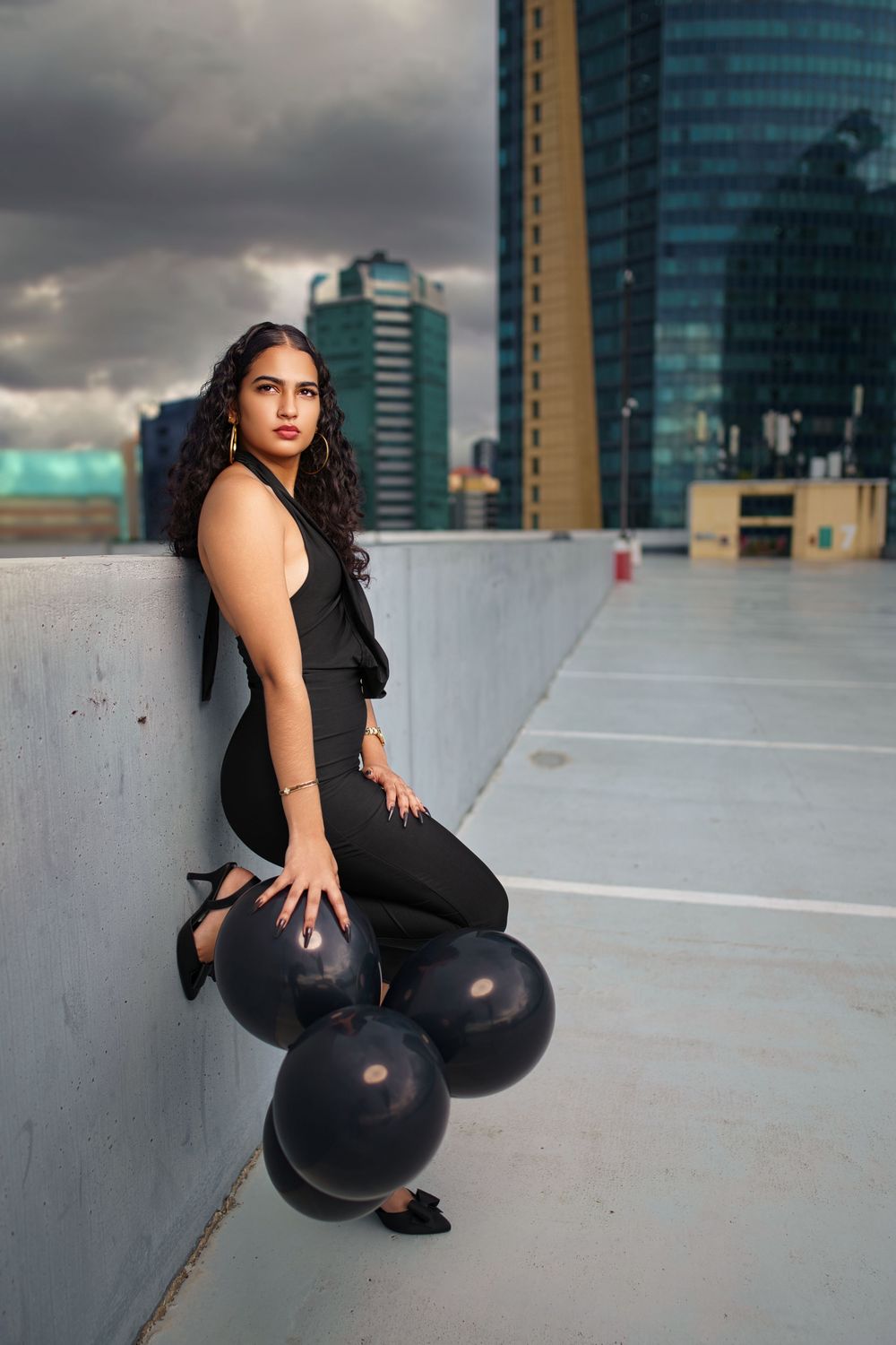 Fashion photoshoot on urban rooftop with black balloons against modern city skyline at sunset.