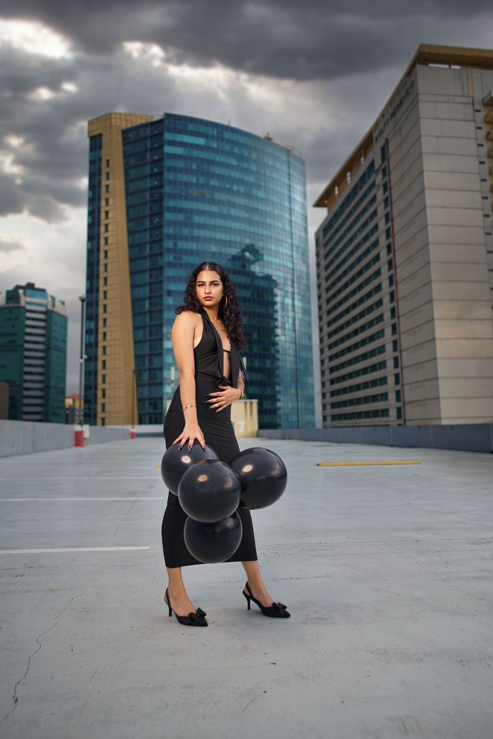 Model in black dress poses with balloons on rooftop against dramatic cityscape with modern glass buildings.