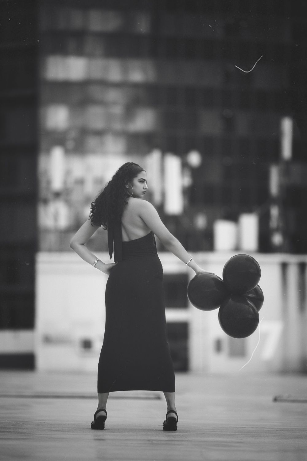 A woman in a black dress holds balloons while posing gracefully in an urban setting, captured in black and white.