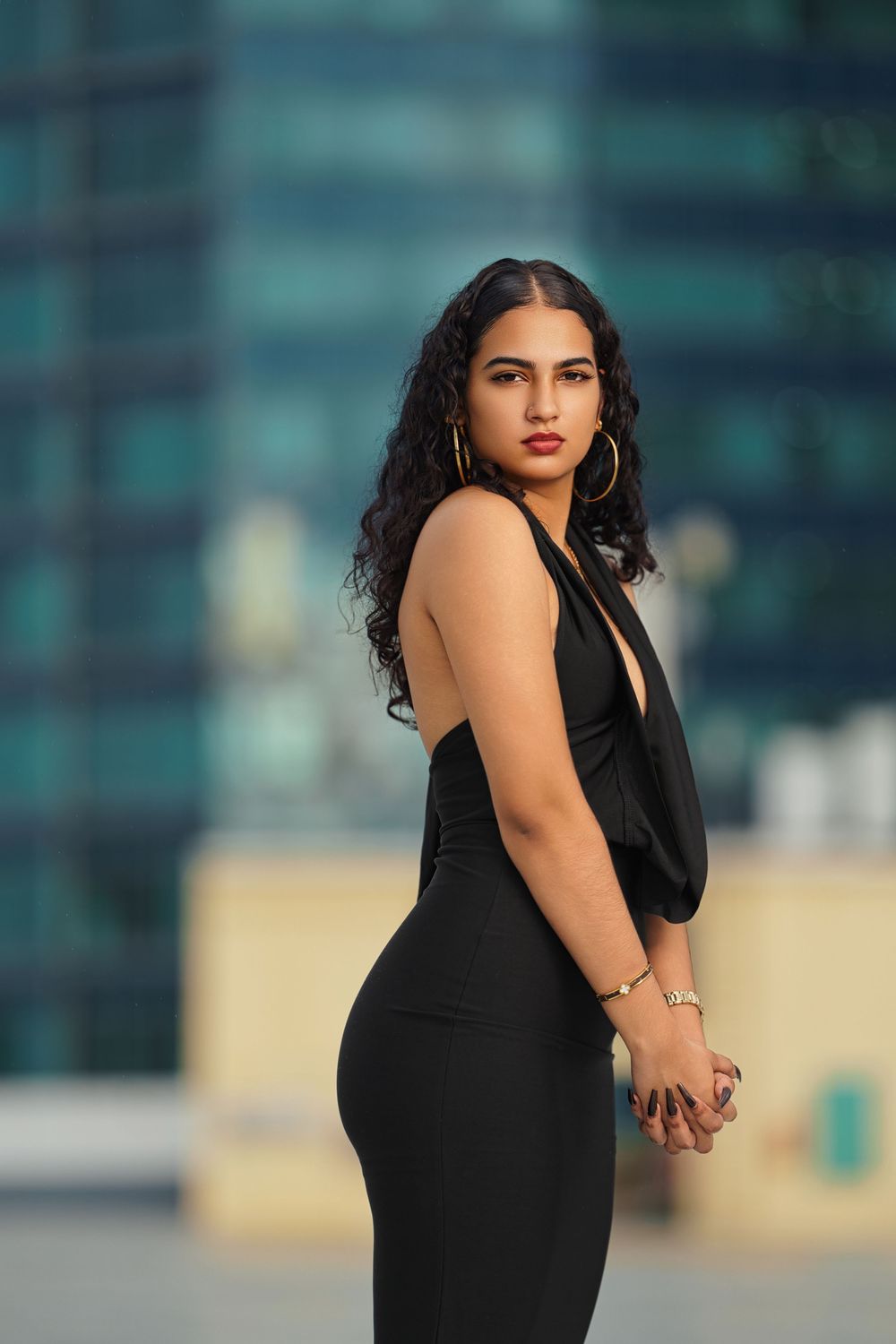 A woman in a black sleeveless dress poses outdoors against a modern architectural backdrop.