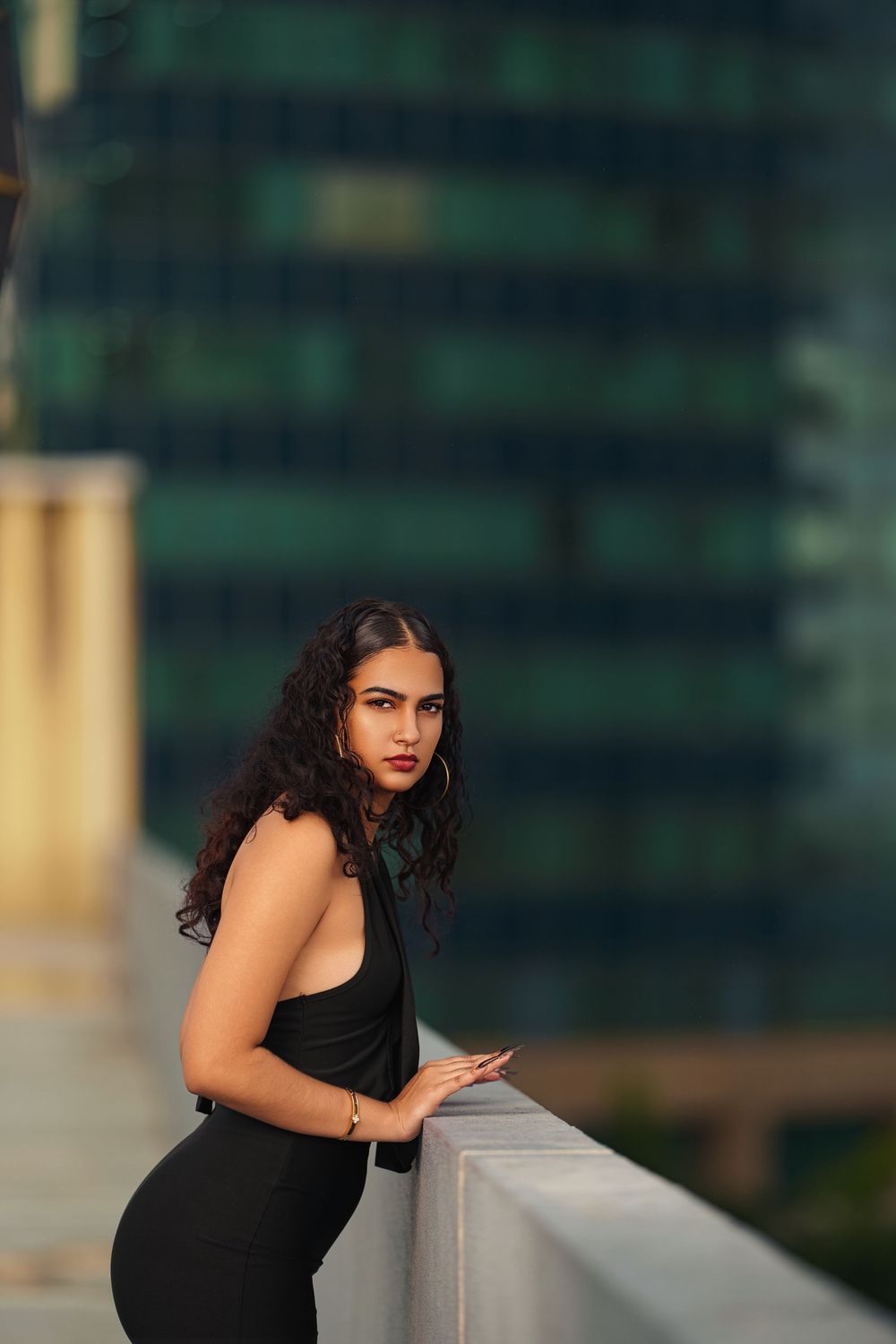 A woman in a black dress leans against a railing with an urban backdrop.