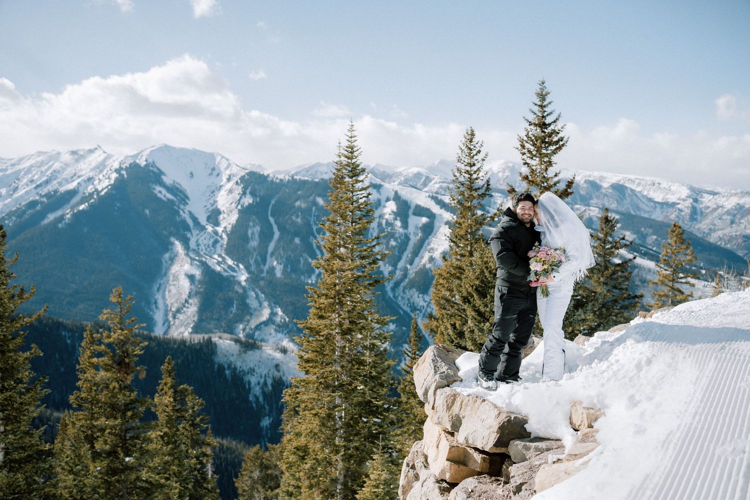 A couple embraces on a mountain peak with evergreen forest and majestic mountain ranges as their backdrop.