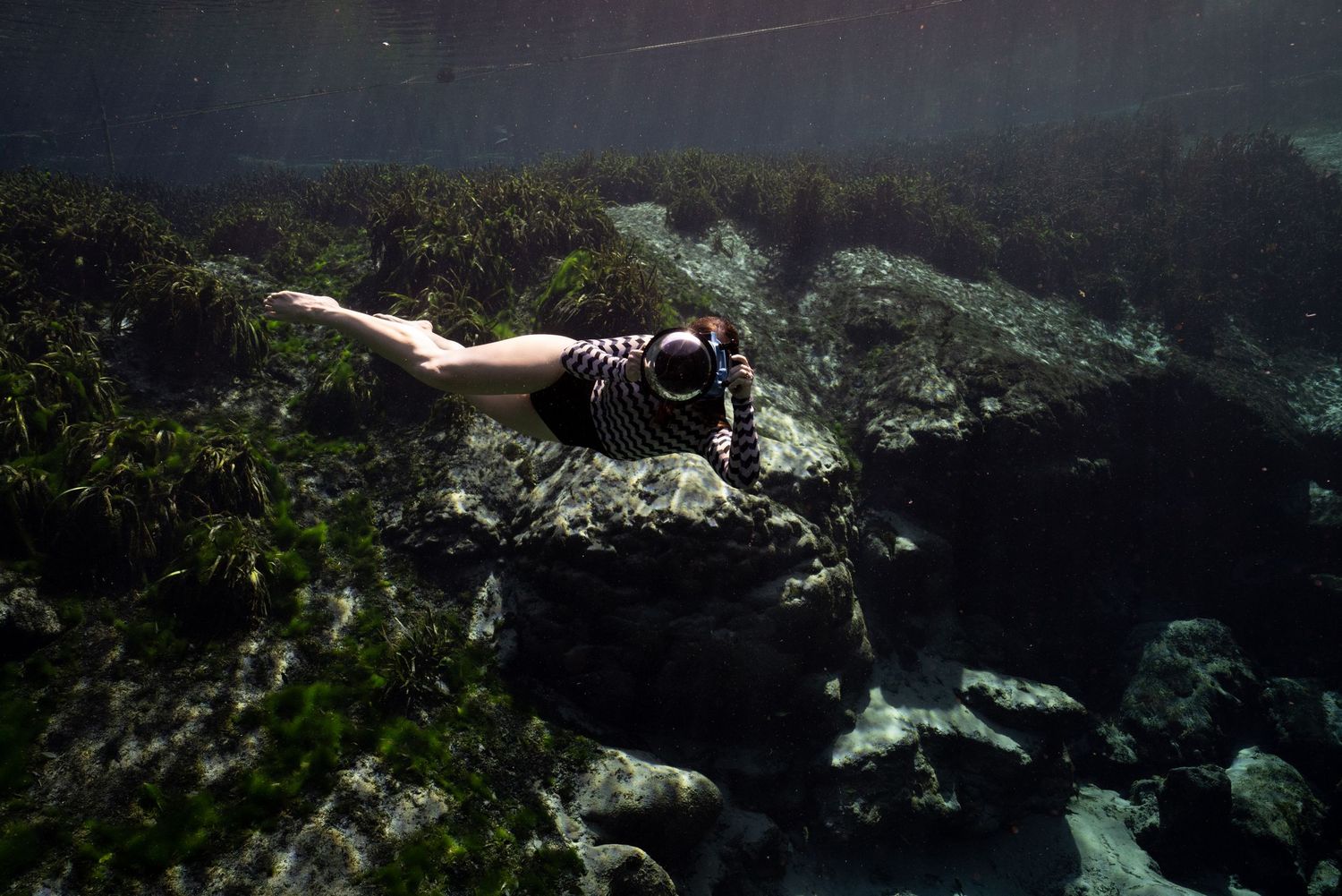A scuba diver swims through clear blue water near coral reefs and rocky underwater formations.