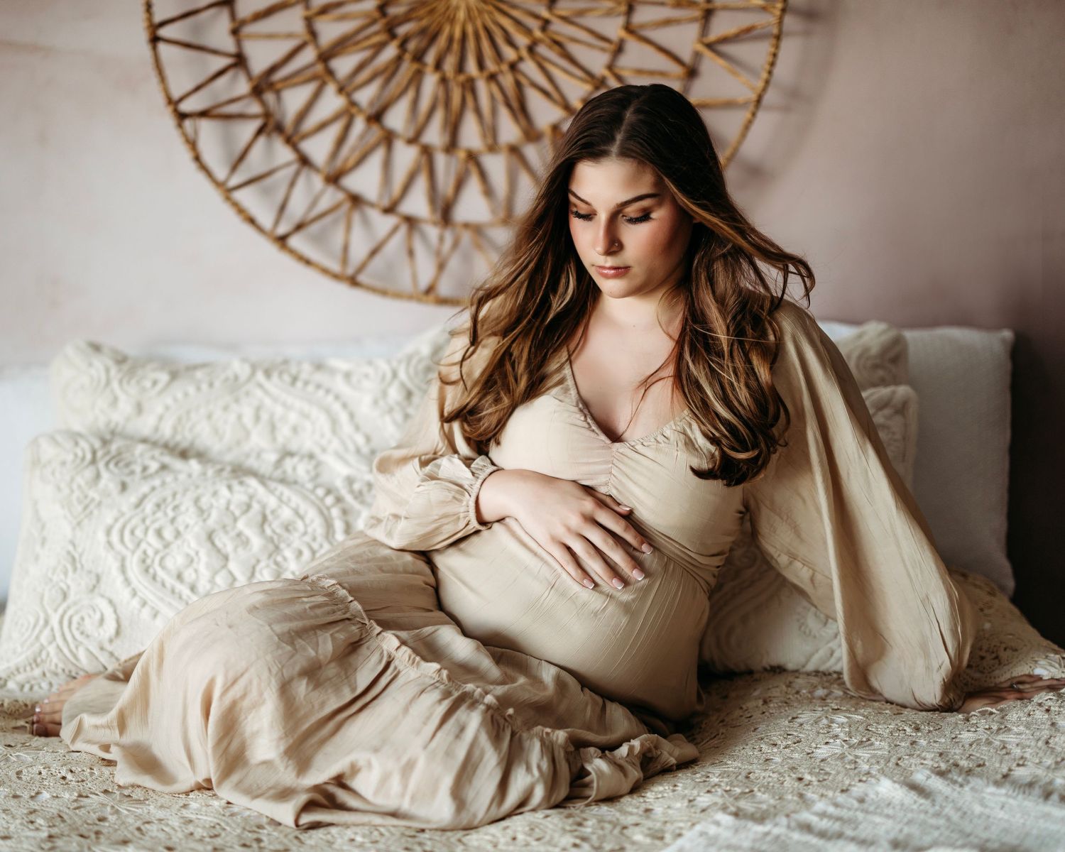 Expectant mother in beige dress sits on white bed beneath decorative circular wall art in neutral tones. Pleasant View studio
