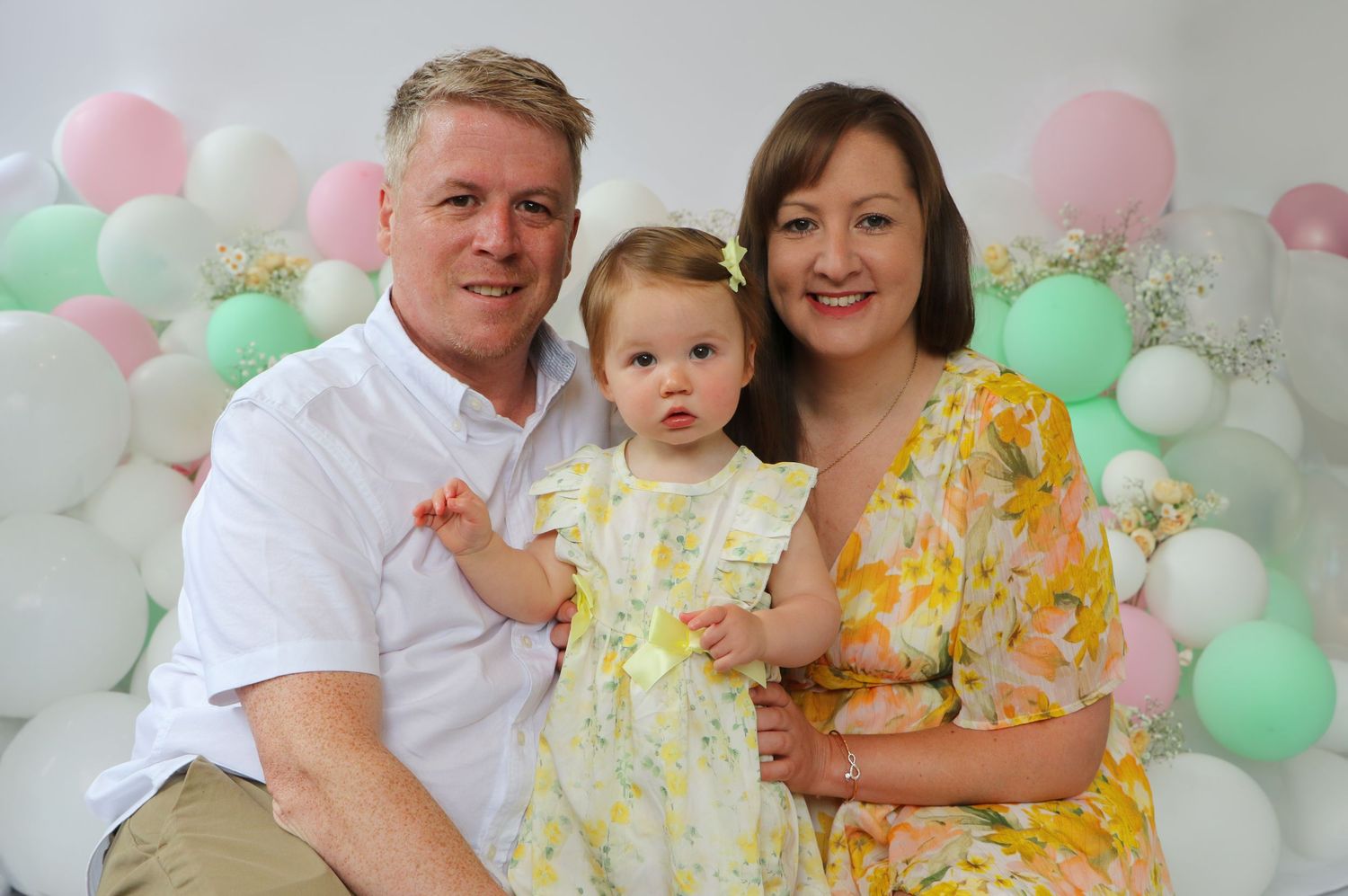 A family poses together at a festive celebration with pink and mint green balloons decorating the background.