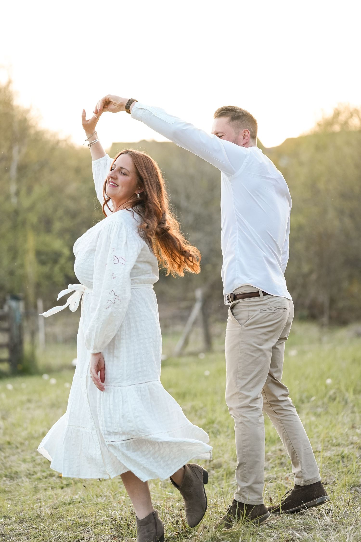 A couple in white and beige outfits dances joyfully together in a sunlit meadow at golden hour.