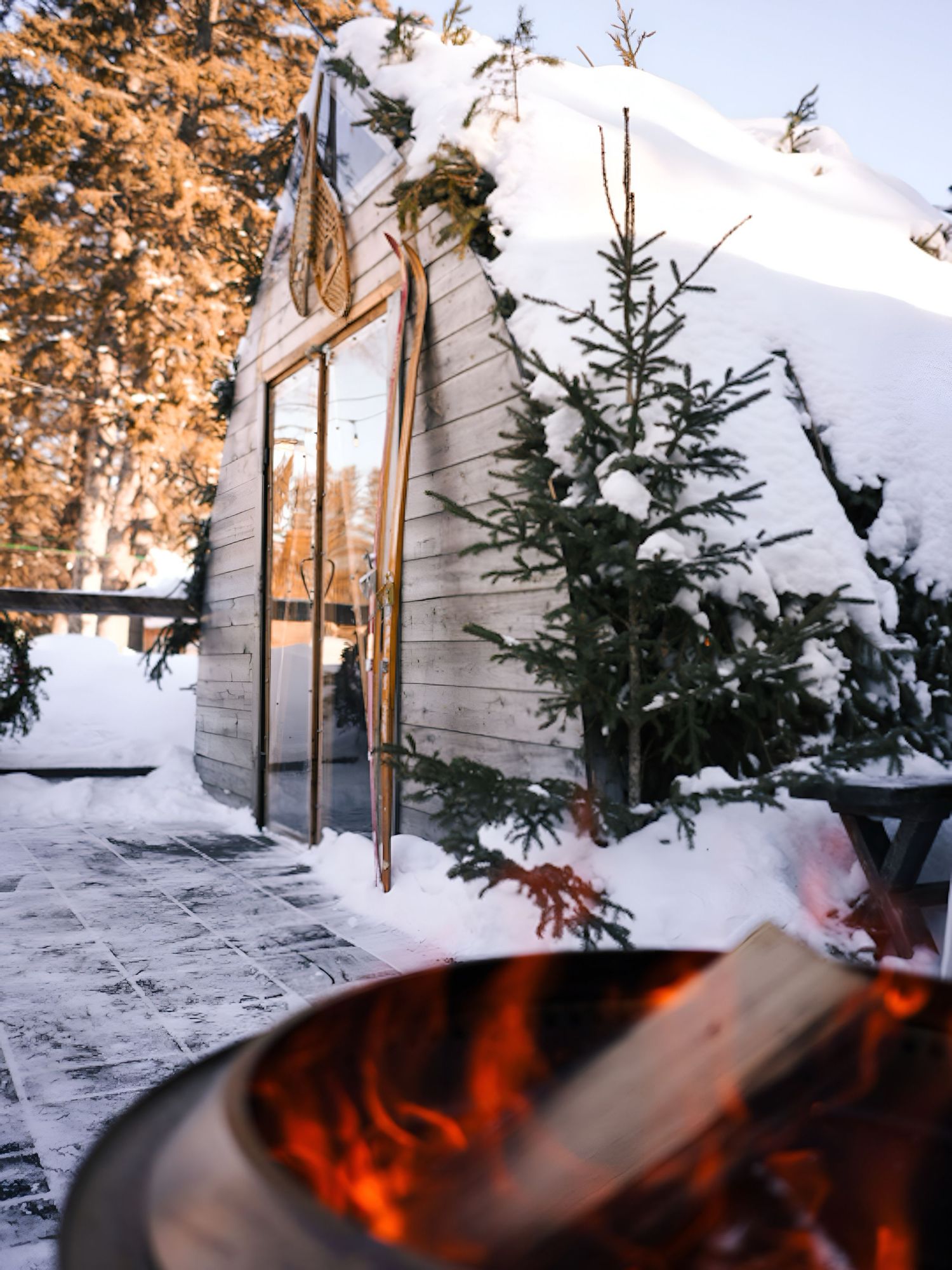 A cozy winter scene with a fire pit glowing in front of a snow-covered cabin in the woods.