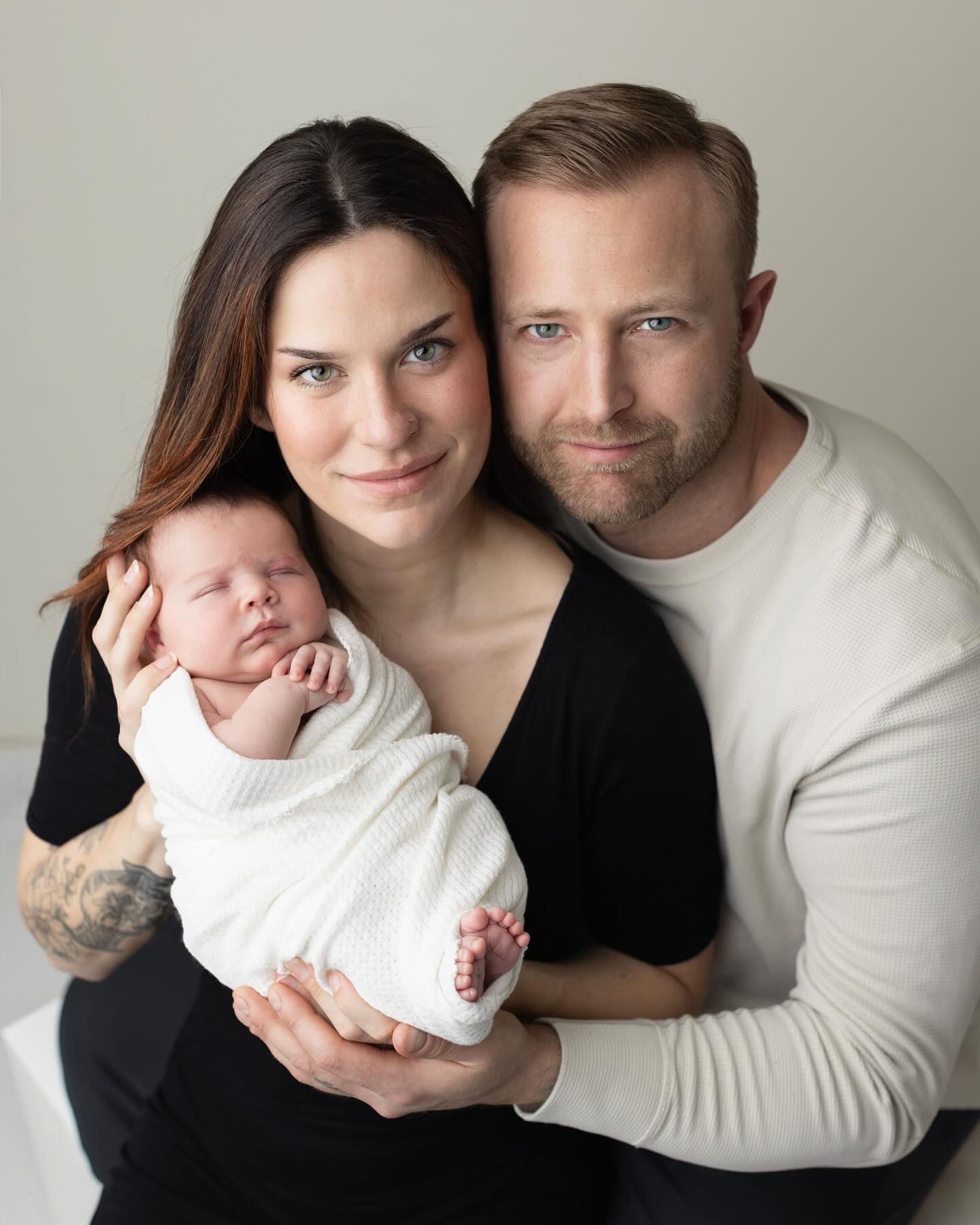 A family portrait featuring parents in black and white clothing holding their sleeping newborn baby in a white outfit.