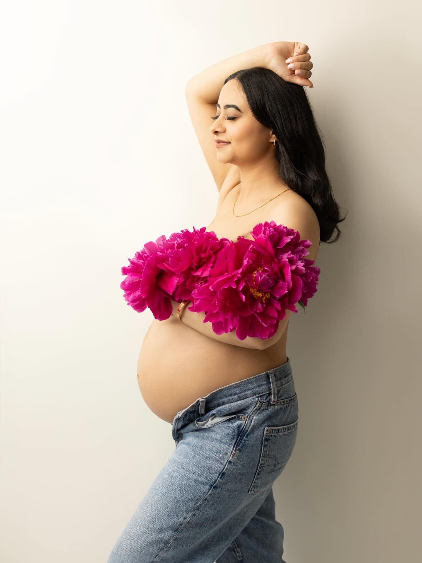 Artistic maternity photo with bright pink peonies and blue jeans against a light background.
