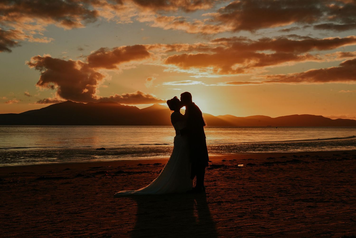 Silhouettes of couples embracing against dramatic orange sunset skies over a beach in West kilbride at Seamill Hydro