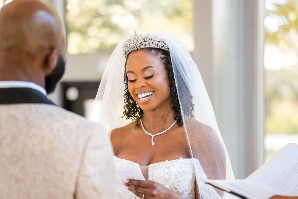 A joyful bride wearing a sparkling tiara and white strapless gown reads from paper during an outdoor wedding ceremony.